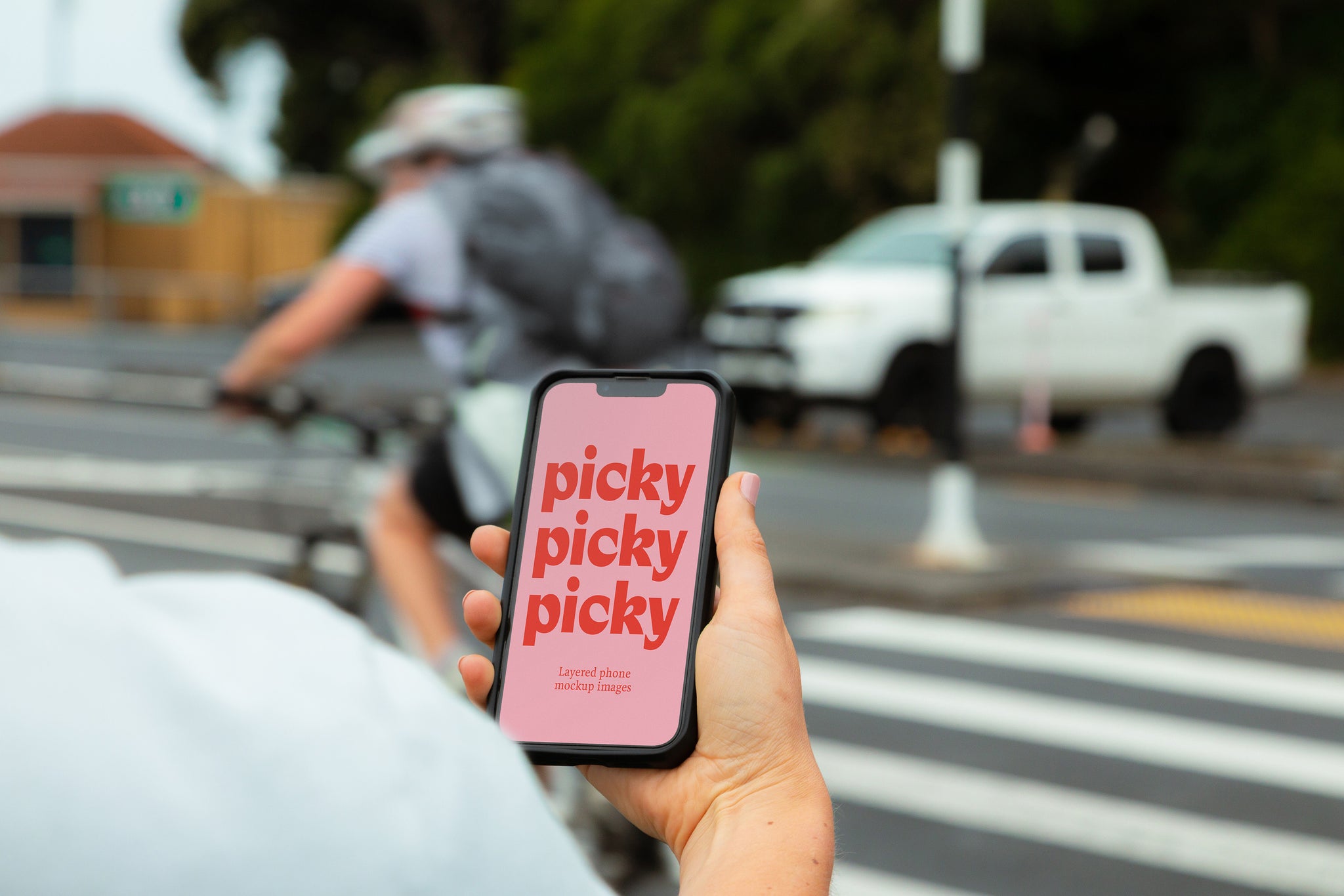 Over shoulder shot of lady holding a smart phone at a pedestrian crossing. Man on bike with backpack in background. Screen facing camera. Bright pink screen with the words 'picky' written three times in red down the phone screen. 