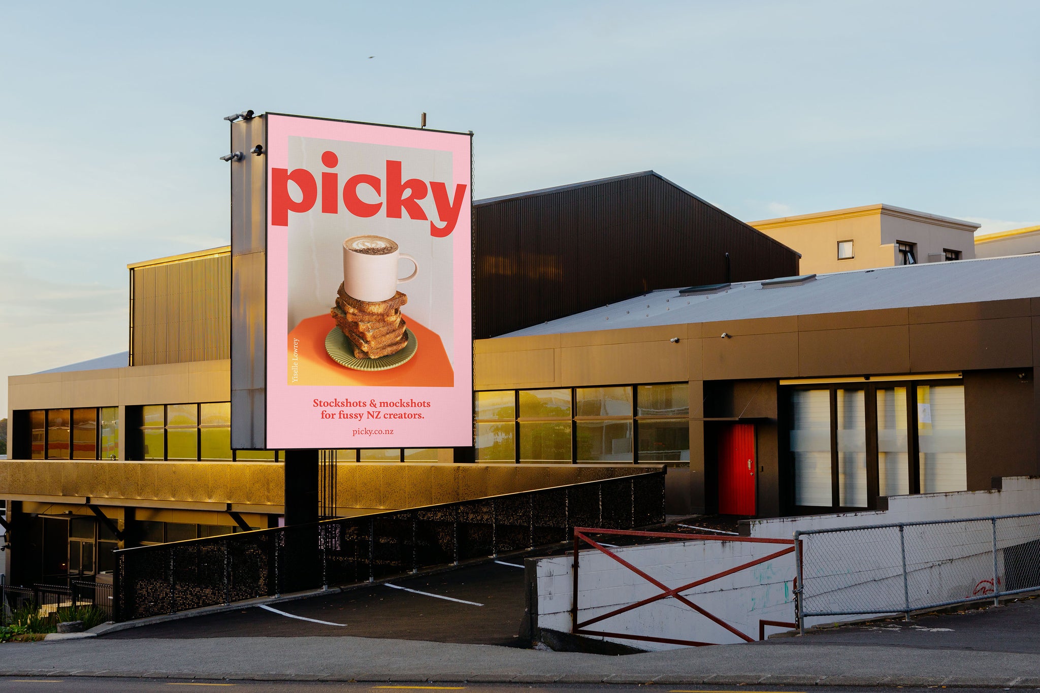 Landscape image of a Auckland building in an urban environment with a large portrait billboard at the front entrance of the building. Soft light, golden hour, blue sky in background. Billboard is pink witha picture and the words 'Picky' written inside. 