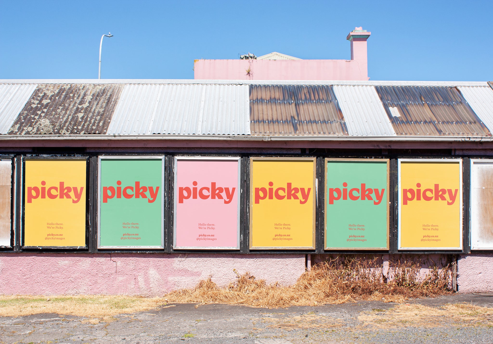 A poster location on a pink building and with a corregated iron roof. Dead weeds grow against this rustic setting.