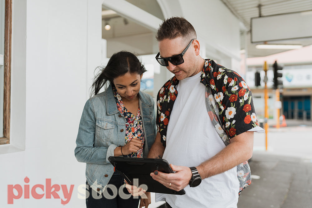 White male in hawaiian shirt and Indian woman in jean jacket look at a black tablet device on a city street.