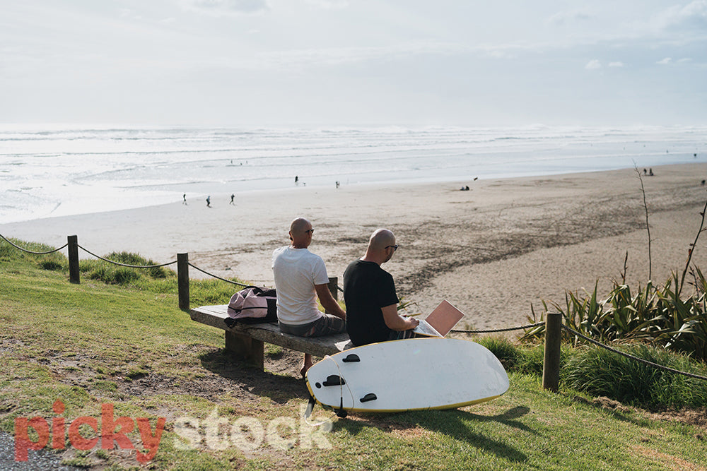 Two men sit on the beach with a surfboard next to them, one using a laptop the other taking a phone call on his mobile phone while the waves break behind them.