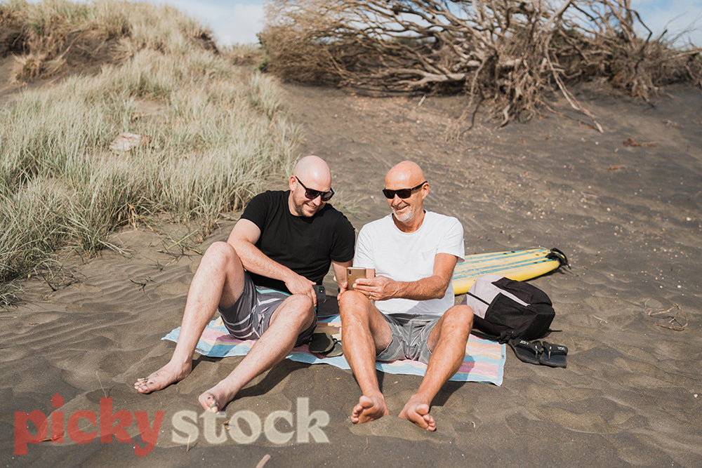Two men in sunglasses and board shorts sit on the beach on a towel one checks their phone.