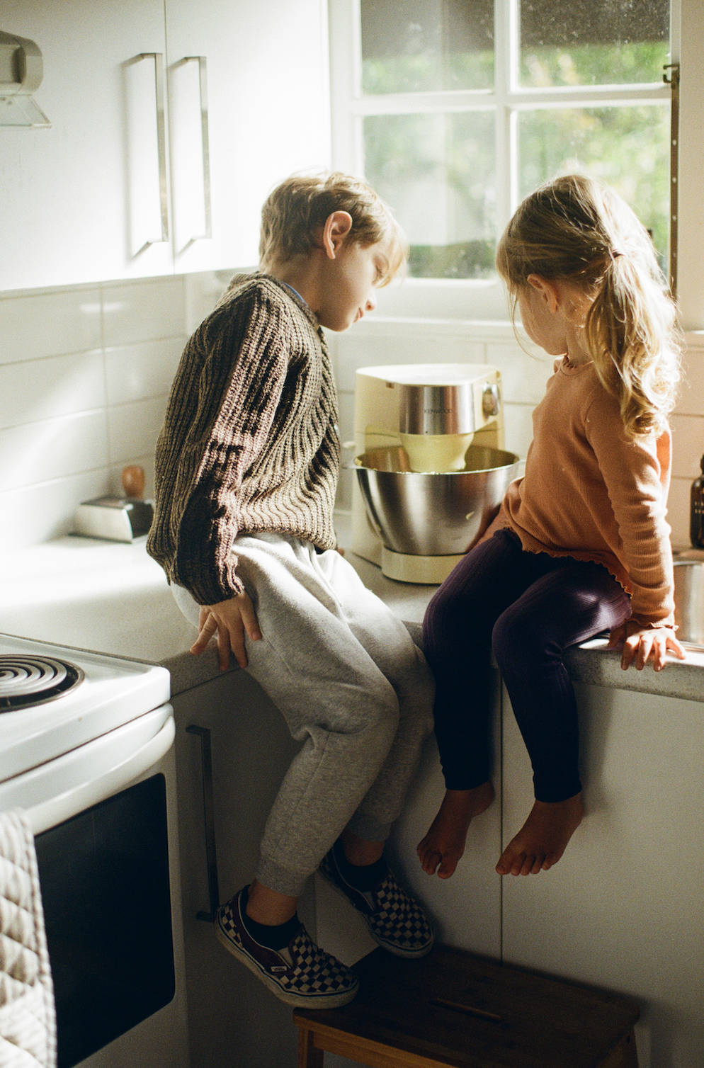 Two young children sit on benchtop corner looking at cakemixer. 