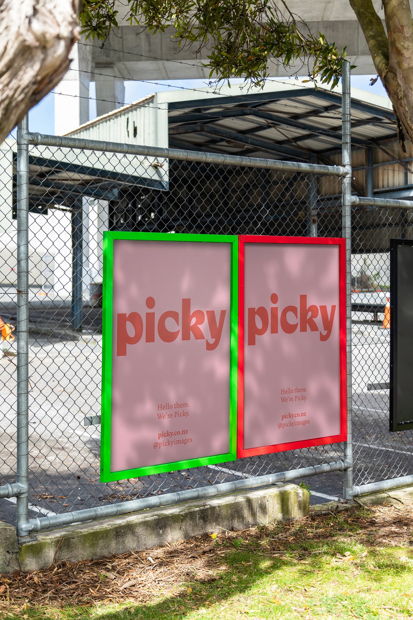 Portrait of colorful advertisement frames on a wire fence sheltered by trees, the frame holds a pink mock advertisement with a red picky logo.
