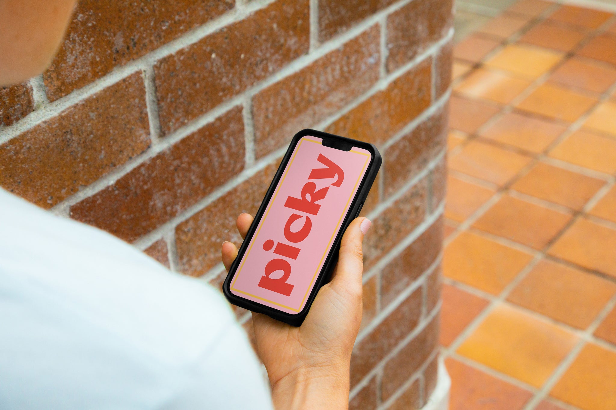 Lady holding smart phone standing against brick wall and path. Smartphone has screen turned on with Picky in red written vertically over pink screen. 