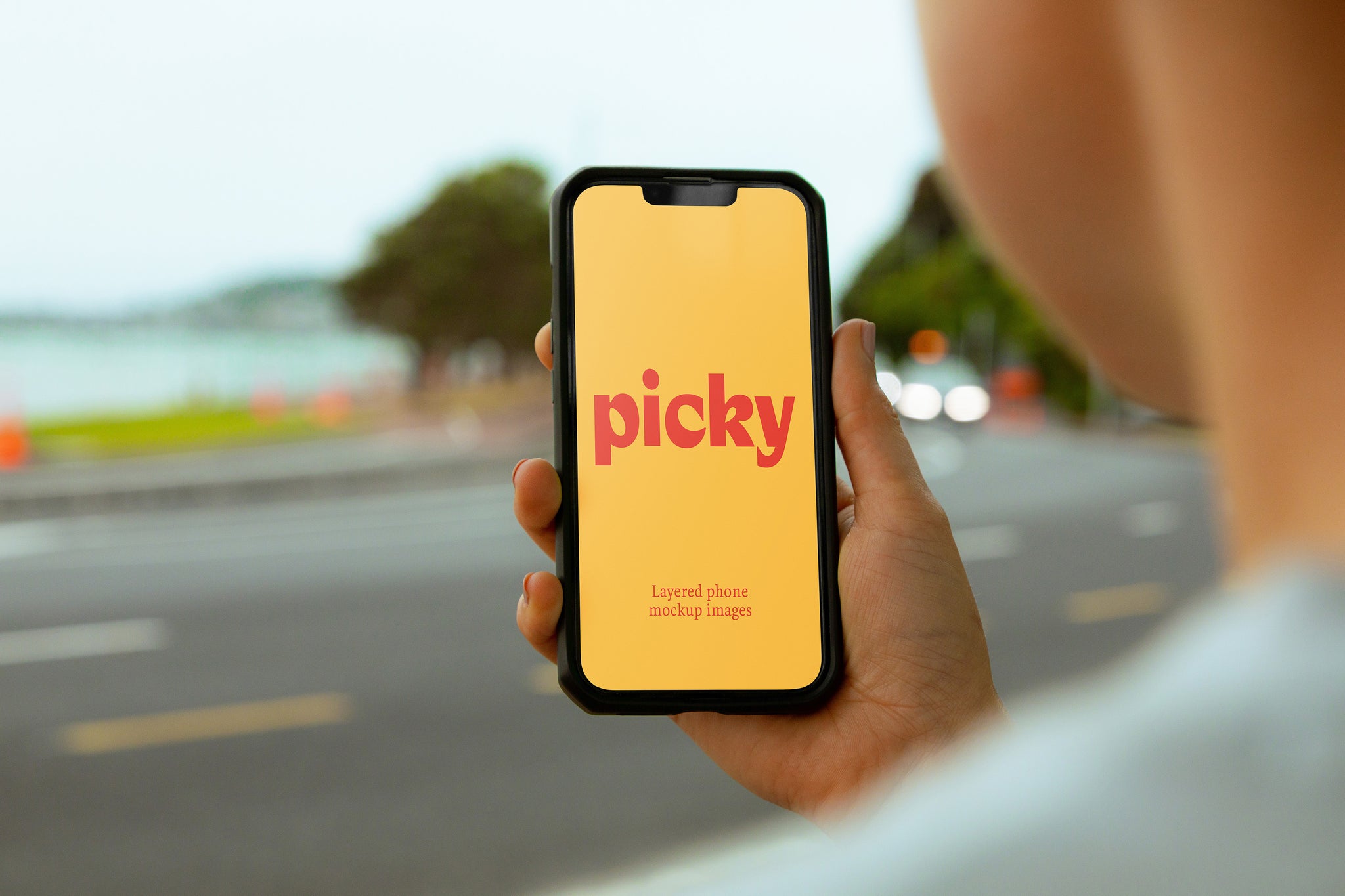 Over the shoulder shot of lady holding phone up right. Picky logo in red written on yellow screen. Background is of a coastal road with ocean in far background. 