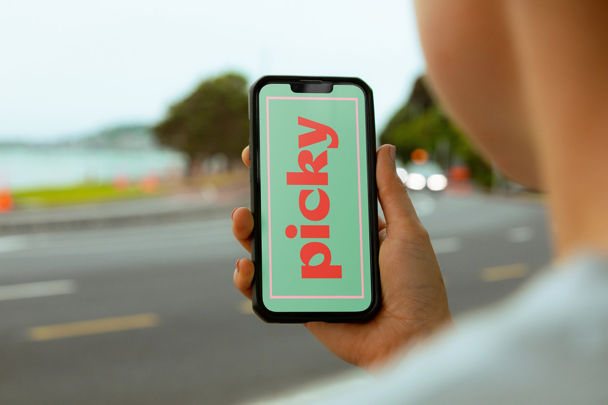 Over the shoulder shot of man holding phone up right. Picky logo vertically doiwn the green screen. Background is of a coastal road with ocean in far background. 