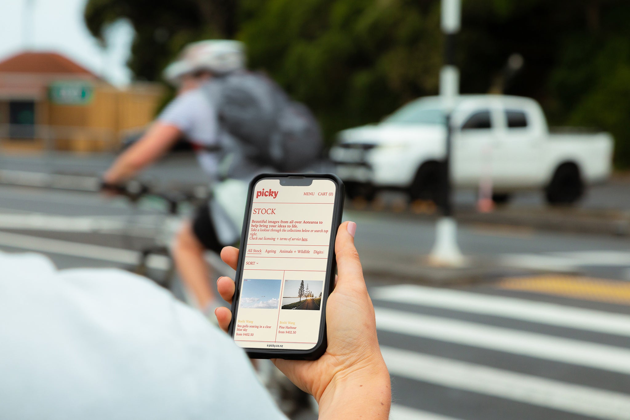Over shoulder shot of man holding a smart phone at a pedestrian crossing. Man on bike with backpack in background. Screen facing camera. Phone screen open on Picky website page. 