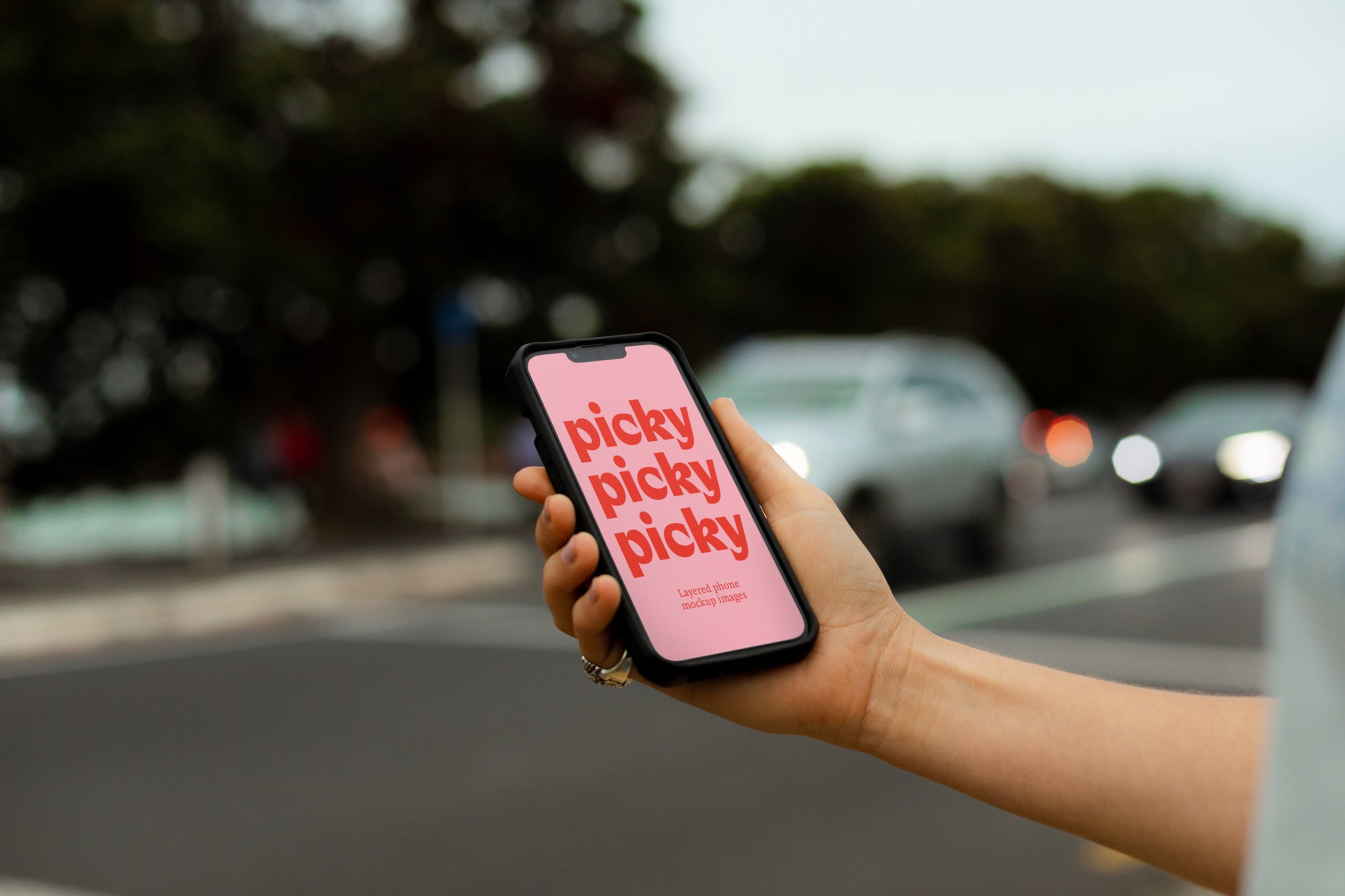 Man holding smart phone, iphone at traffic light crossing. Holding phone out, body cropped. Cars with headlights visible. Bright pink screen with the words 'picky' written three times in red down the phone screen. 