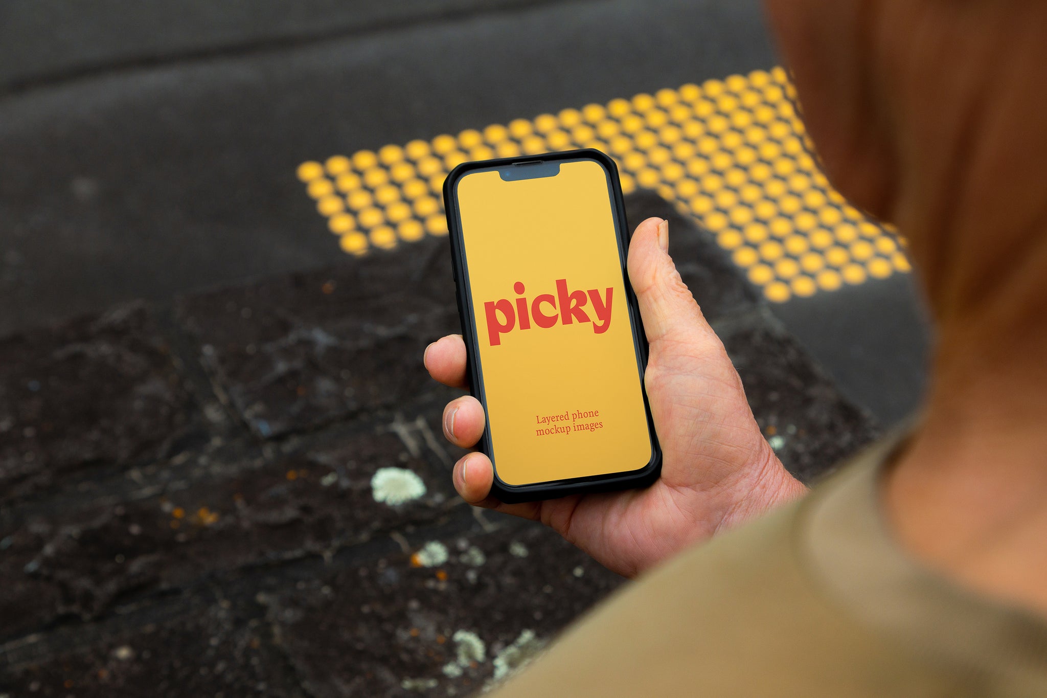 Man holding smart phone, iphone at traffic light crossing. Over the shoulder image with yellow crossing markers on the black concrete. Bright yellow screen with the words 'picky' written in red. 