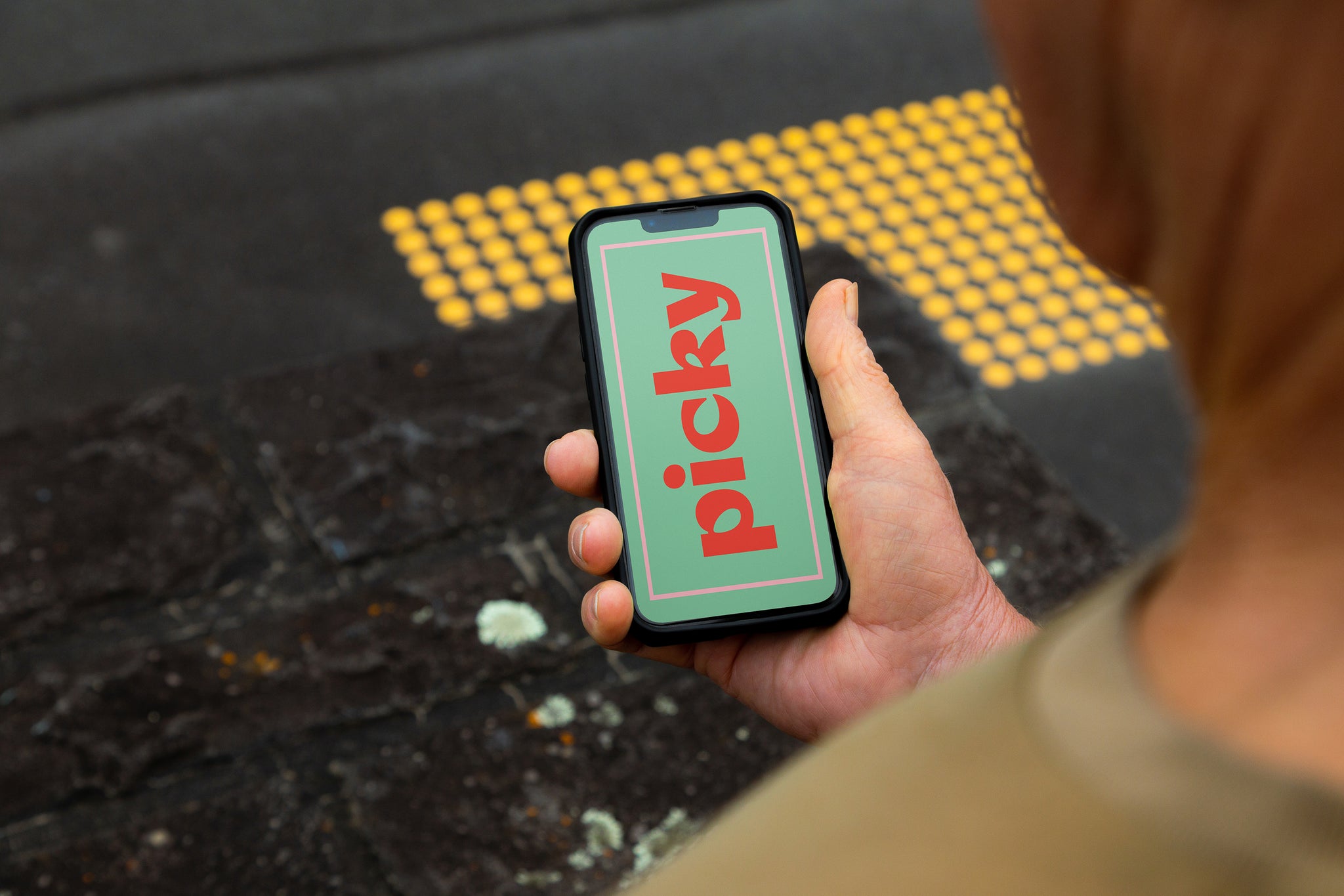 Man holding smart phone, iphone at traffic light crossing. Over the shoulder image with yellow crossing markers on the black concrete. Bright green screen with the words 'picky' written vertically in red. 
