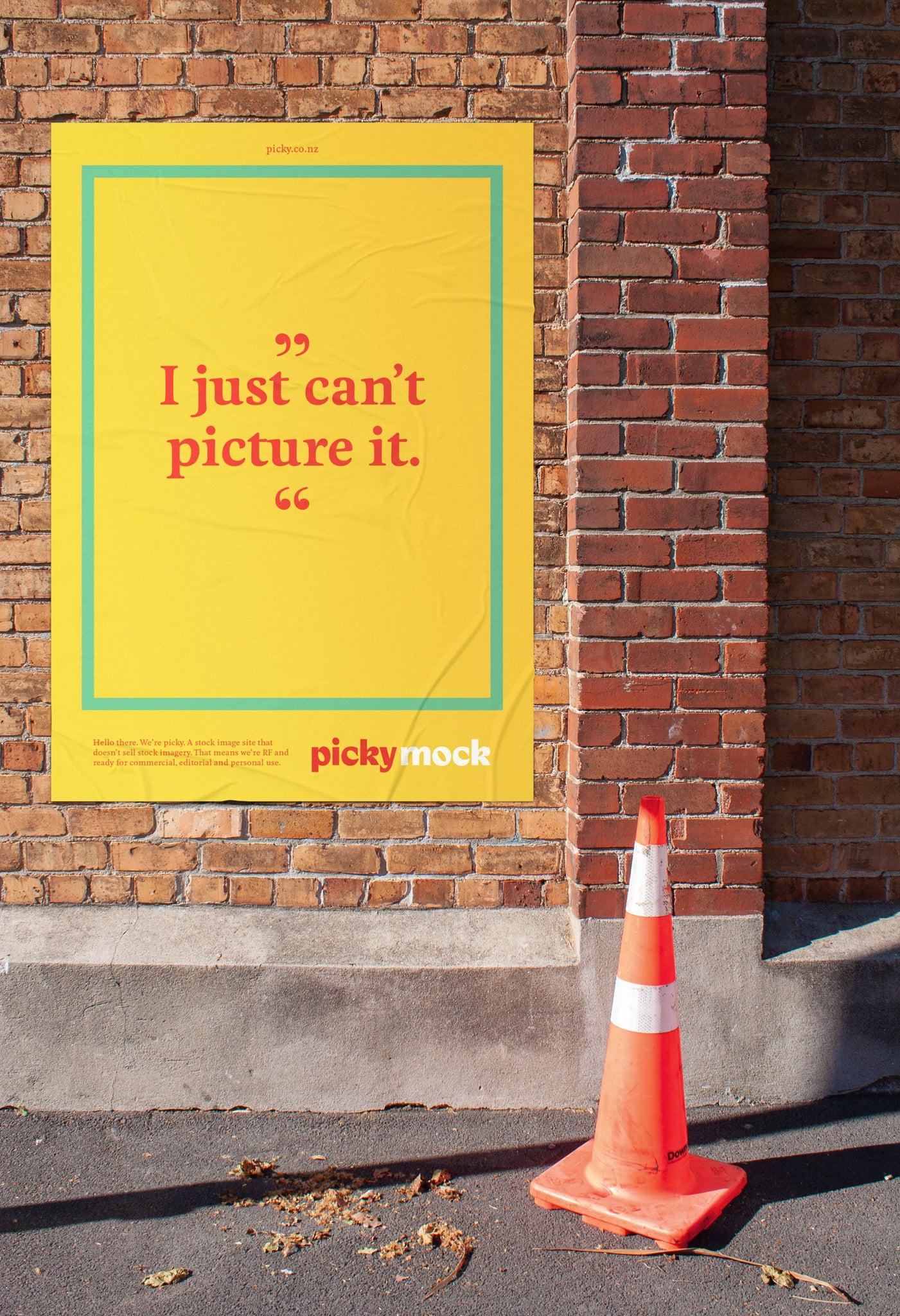 A mockup image of a large portrait street poster set against a red brick urban wall. A bright orange road cone sits to the right of the poster on the footpath. The poster showcases placeholder Picky images branding. 