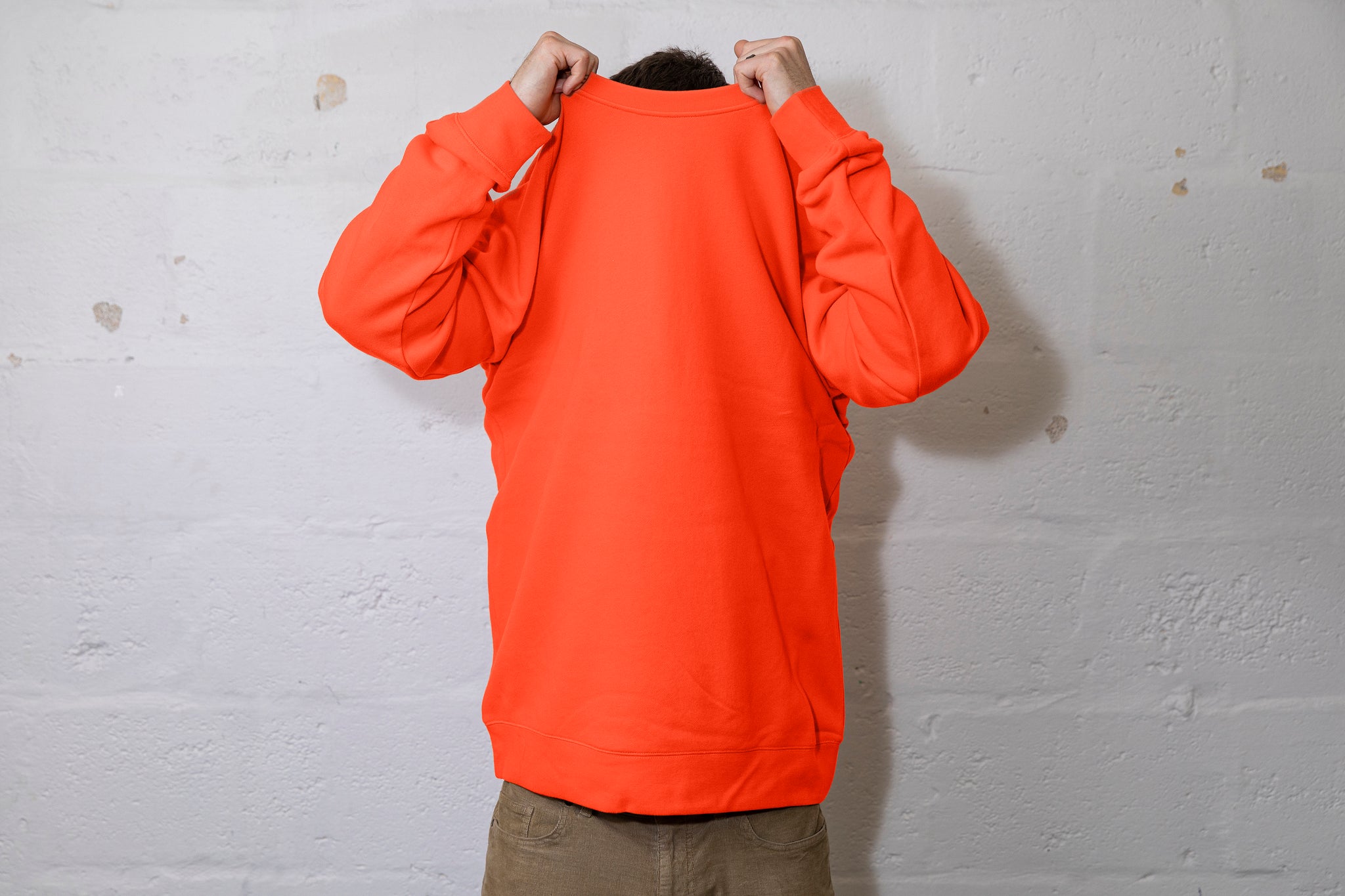 Person pulling a large orange jumper over their head. Can see the top of their dark hair. White cinderbock background. 