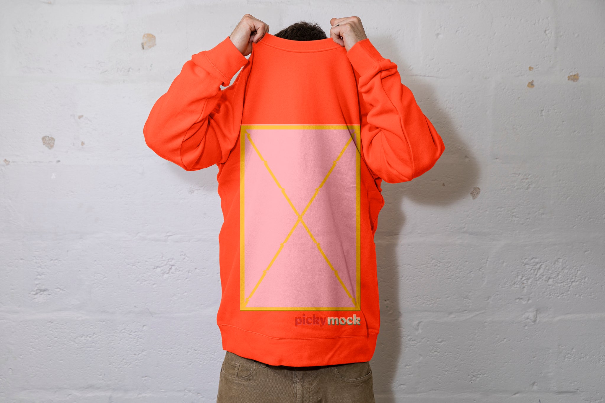 Person pulling a large orange jumper over their head. Can see the top of their dark hair. Pink box in the middle of the jersey with a yellow outline. Picky Mock written under the bottom left of the box. White cinderbock background. 