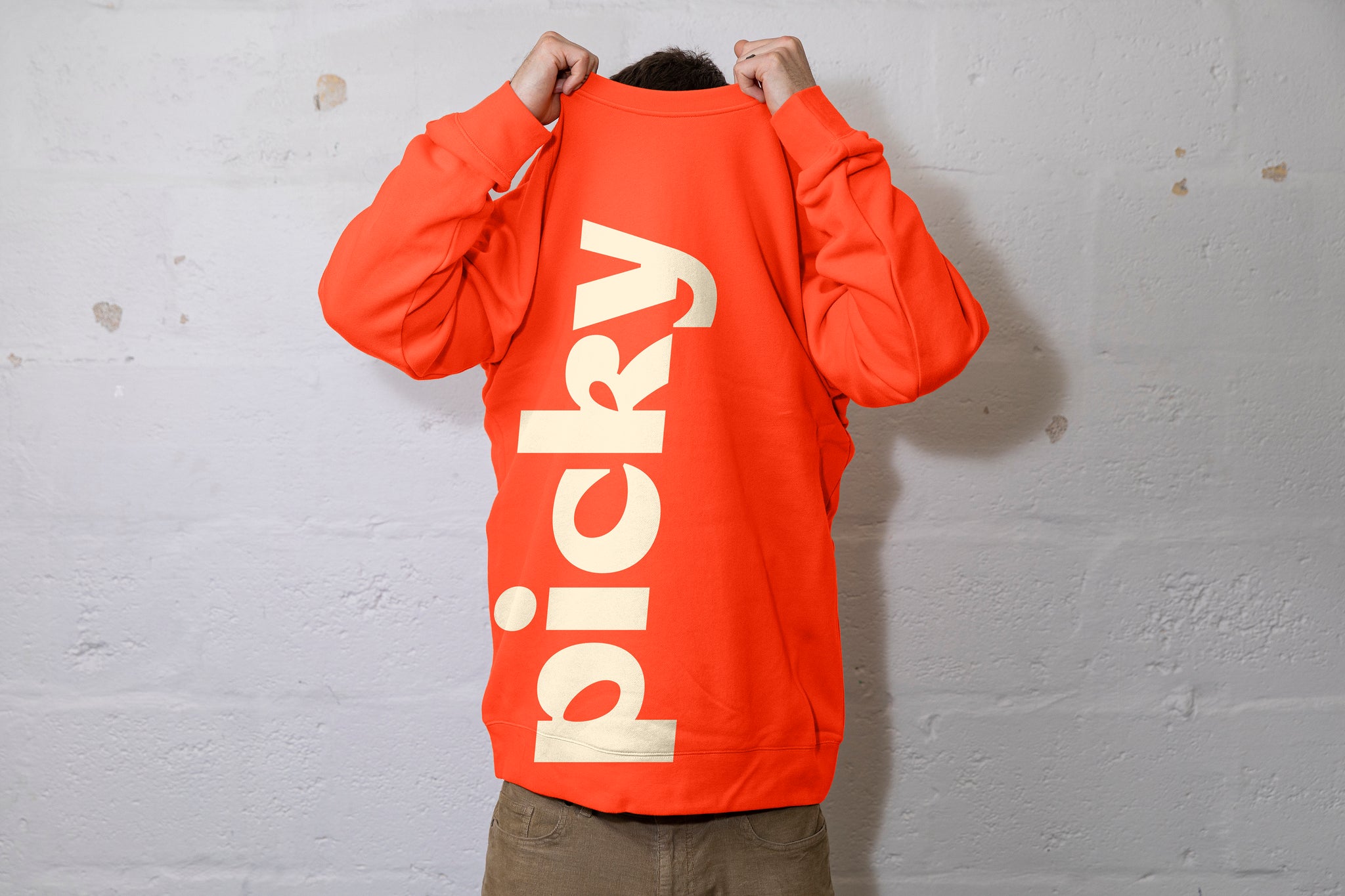 Person pulling a large orange jumper over their head. Can see the top of their dark hair. The word Picky is written in a bold font vertically up the jersey. White cinderbock background. 