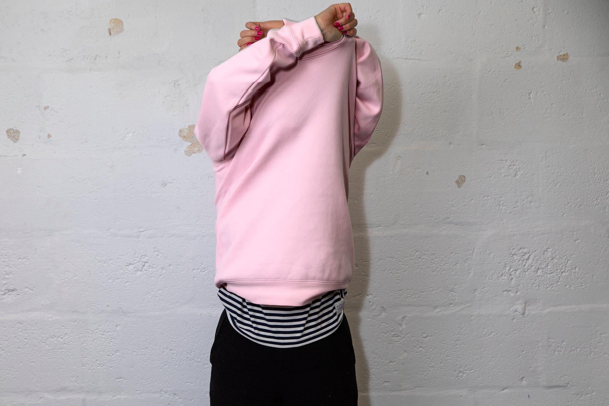 Person pulling a large pink jumper over their head with both hands above their head. White cinderbock background. 