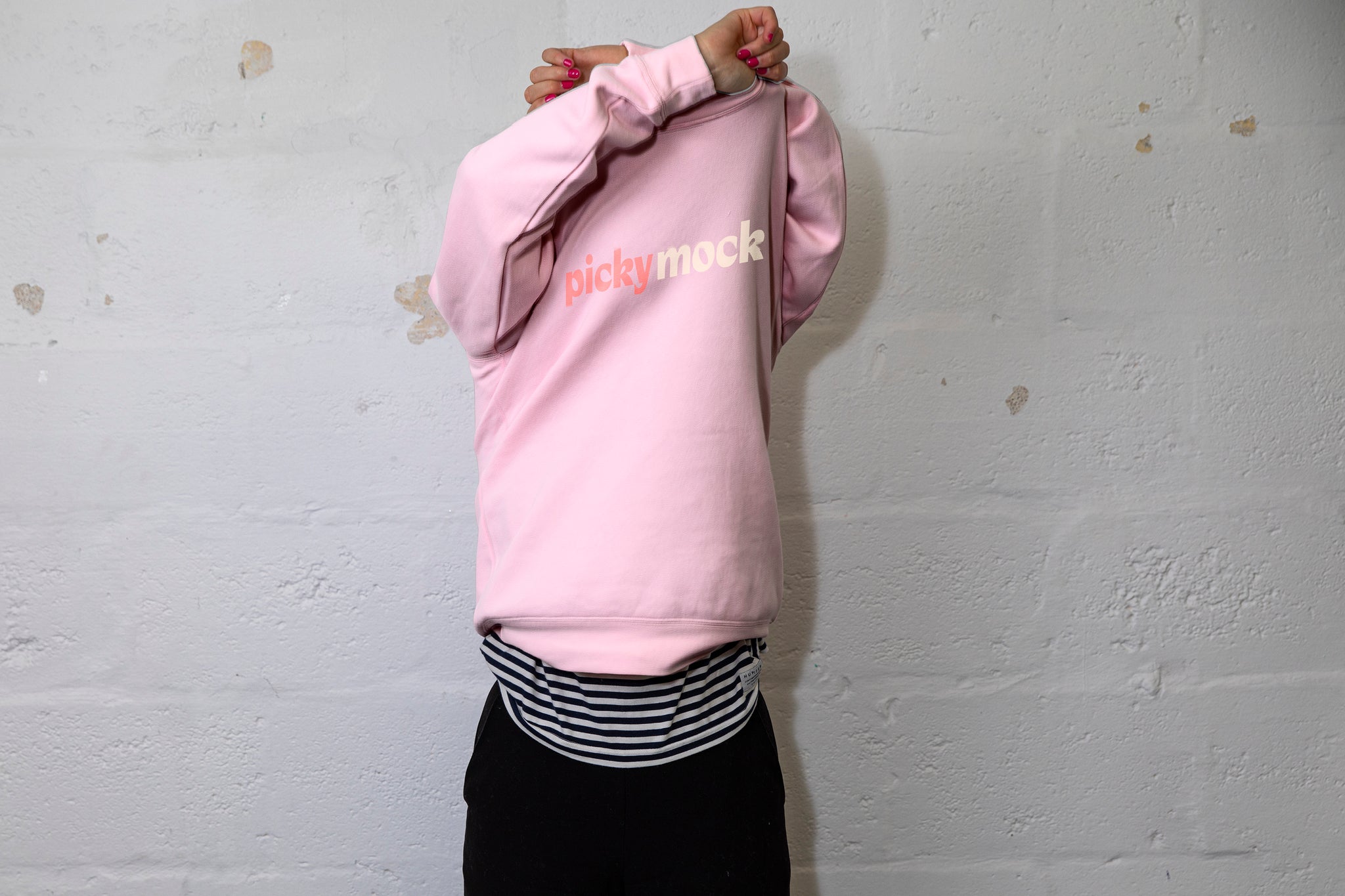 Person pulling a large pink jumper over their head with both hands above their head. Picky Stock written across the chest. White cinderbock background. 