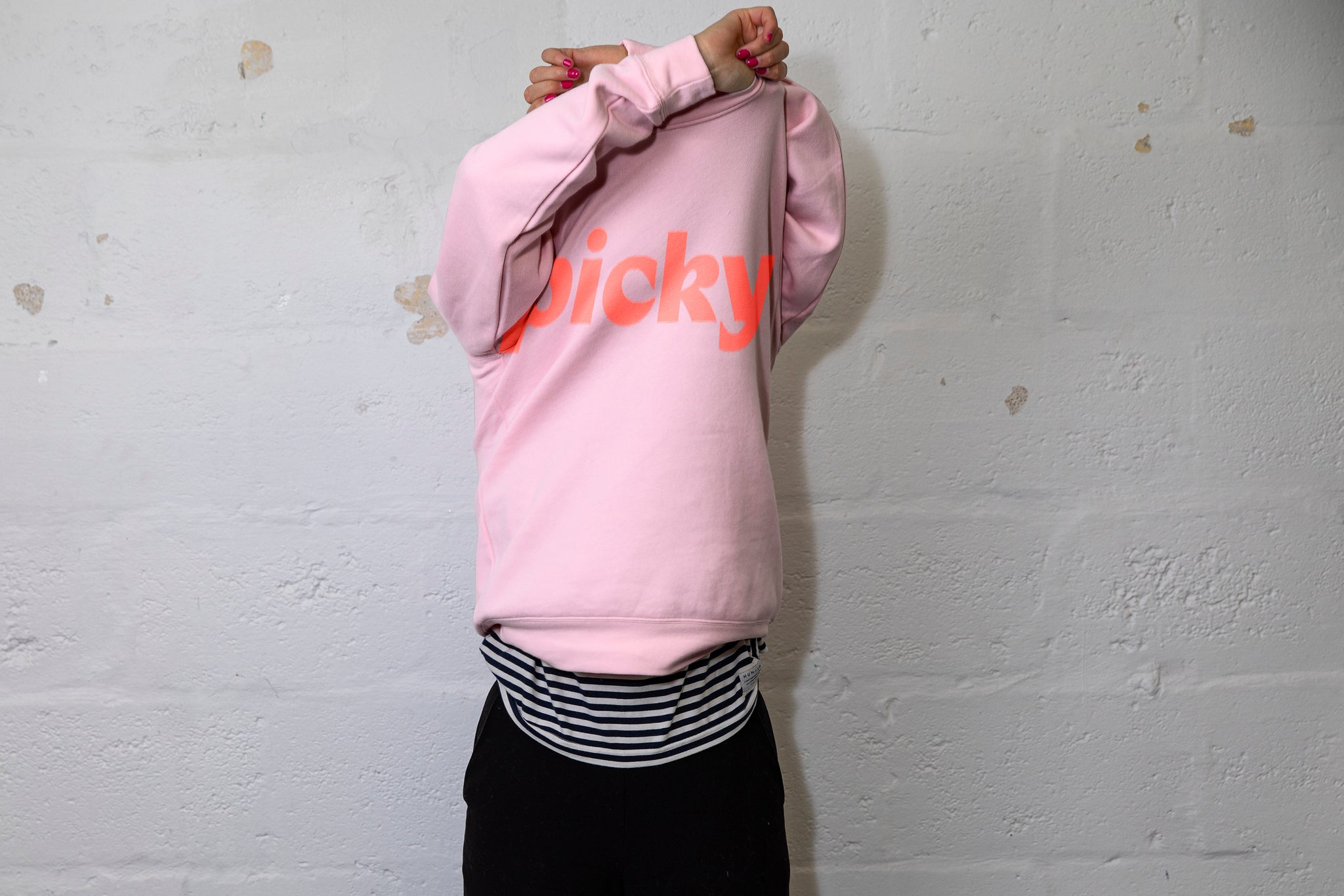 Person pulling a large pink jumper over their head with both hands above their head. Picky Stock in red written in a large font across the chest. White cinderbock background. 