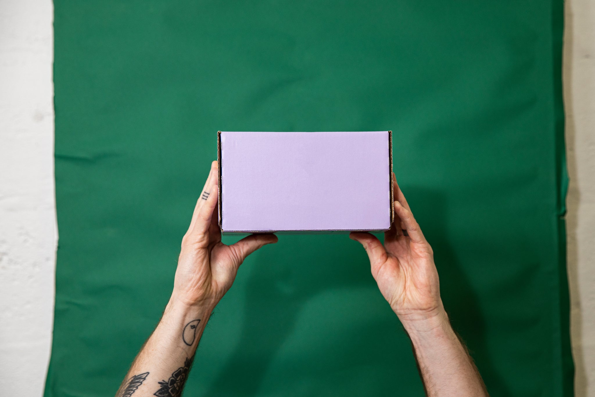 Tattooed hand holding up a box of cans. Green fabric background. The box is purple. 