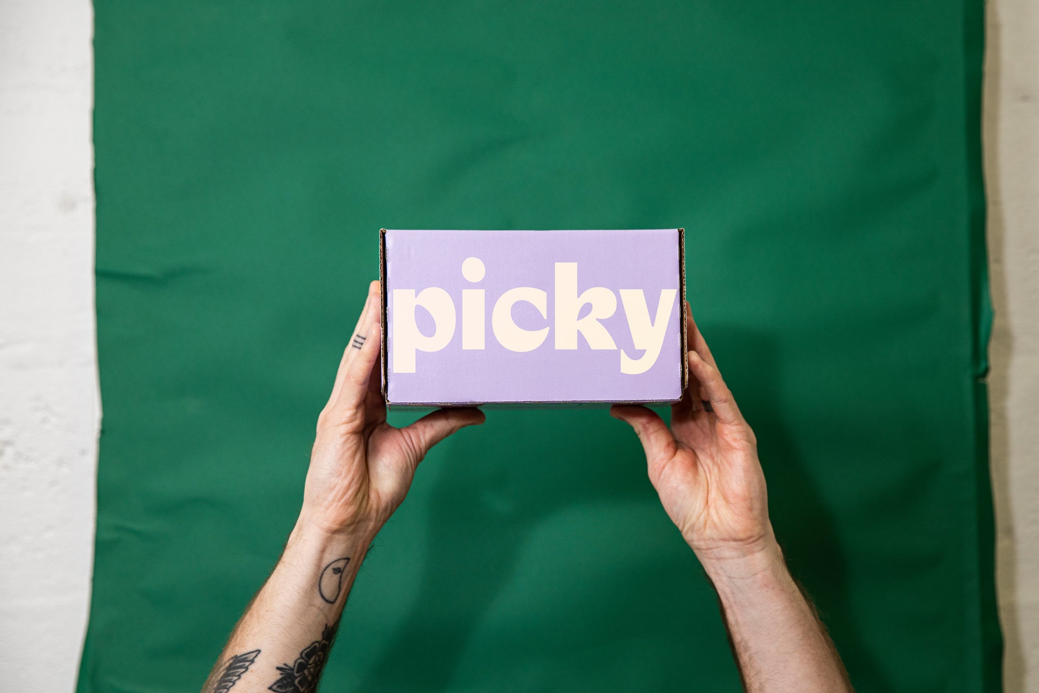Tattooed hand holding up a box of cans. Green fabric background. The box is purple with the words Picky written on the front in a large white font.  