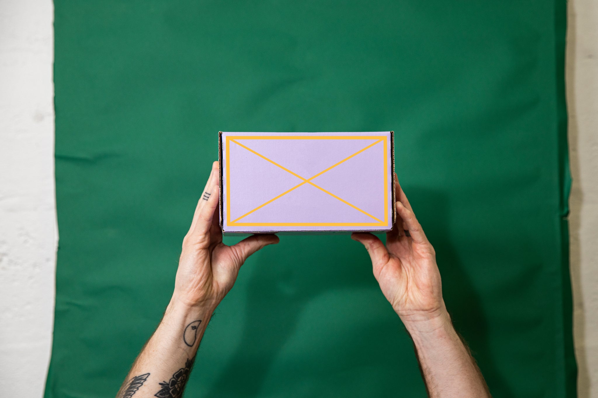 Tattooed hand holding up a box of cans. Green fabric background. The box is purple. With a pink grid line around the front of the box with a X in the middle. 
