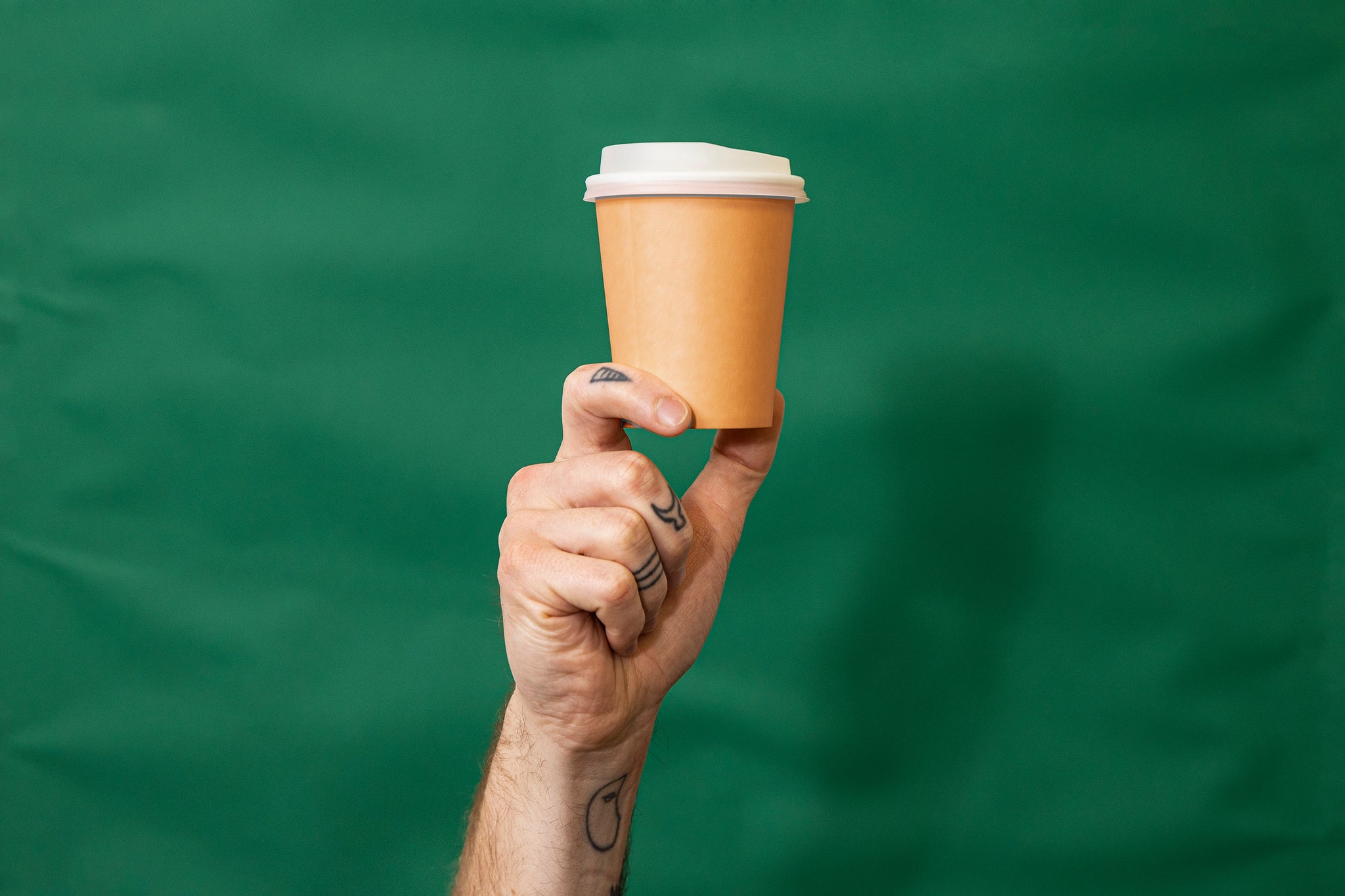 Tattooed hand holding up an orange disposable coffee cup with white lid. Green fabric background. 