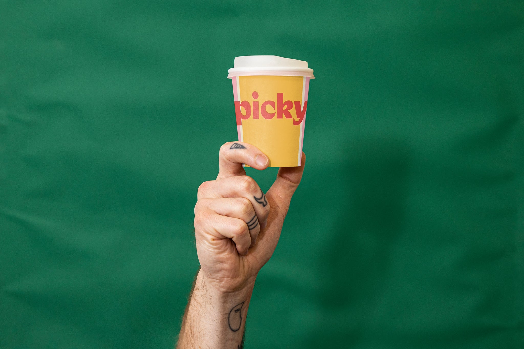 Tattooed hand holding up an orange disposable coffee cup with white lid. The word Picky is written in red on the front of the cup. Green fabric background. 