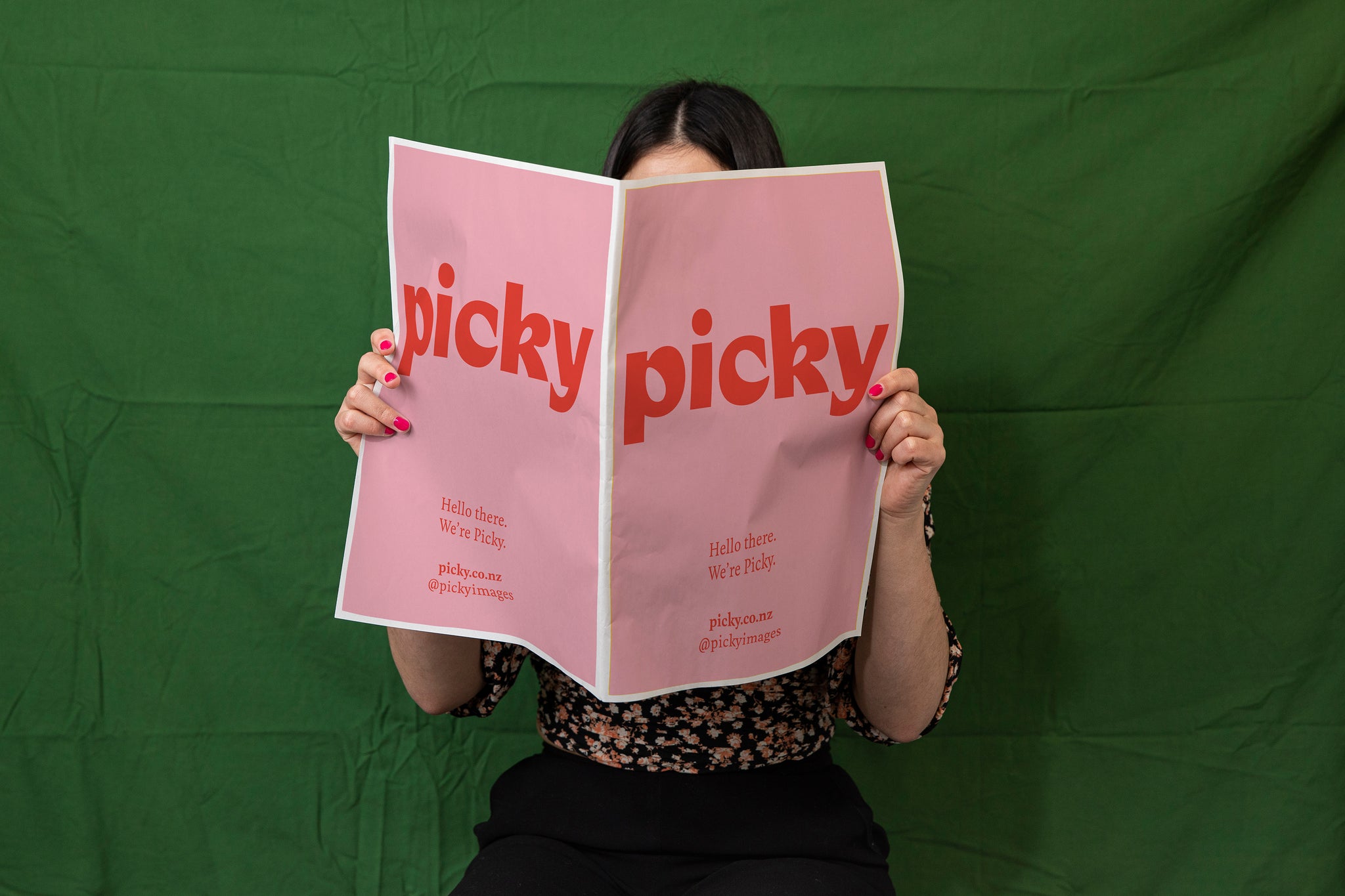Landscape close-up of a woman in pink nail polish reading a large paper document in front of a green backdrop; it is pink with a large red picky logo and mock-up advertisement.