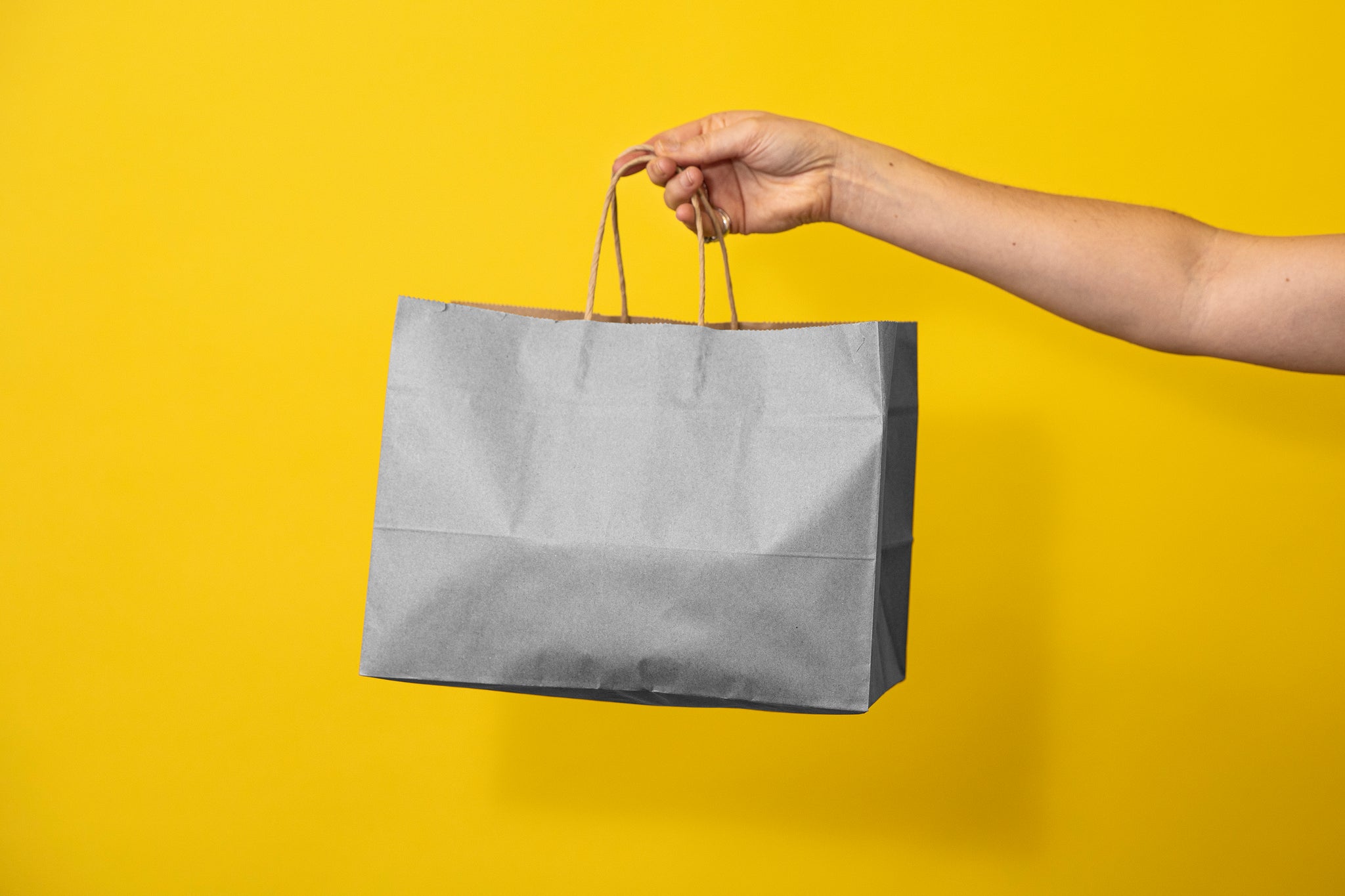 Woman's arm holding out a plain bag to the middle of the frame Background is a bright yellow. 