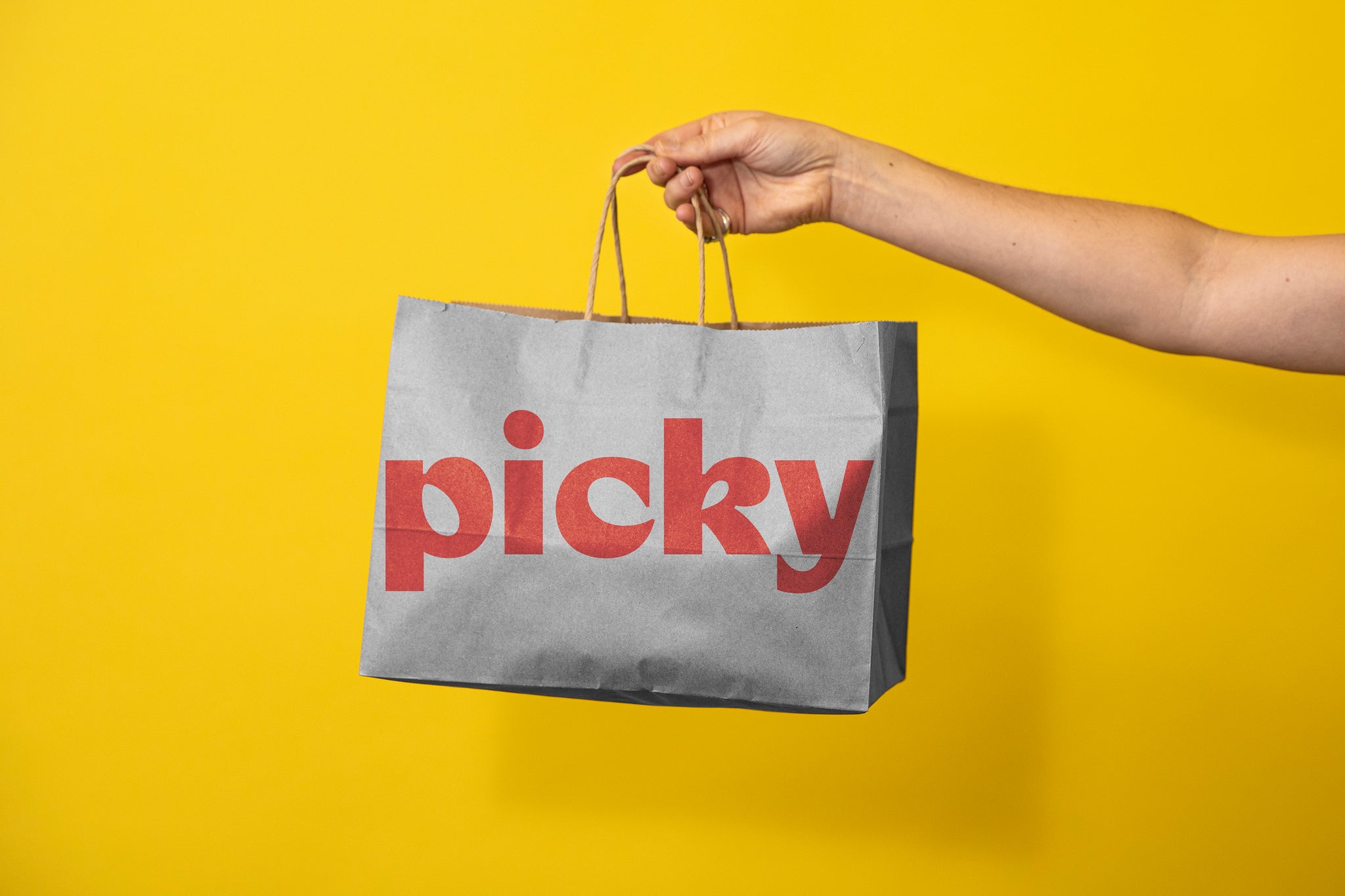 Woman's arm holding out a paper bag to the middle of the frame. The word Picky is written in red, largely across the front. Background is a bright yellow. 
