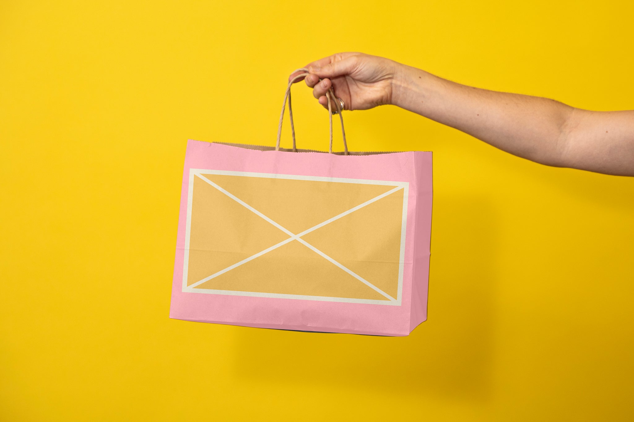 Woman's arm holding out a pink paper bag to the middle of the frame. Orange box with a white border grid line with an X in the middle of the bags desgin. Background is a bright yellow. 