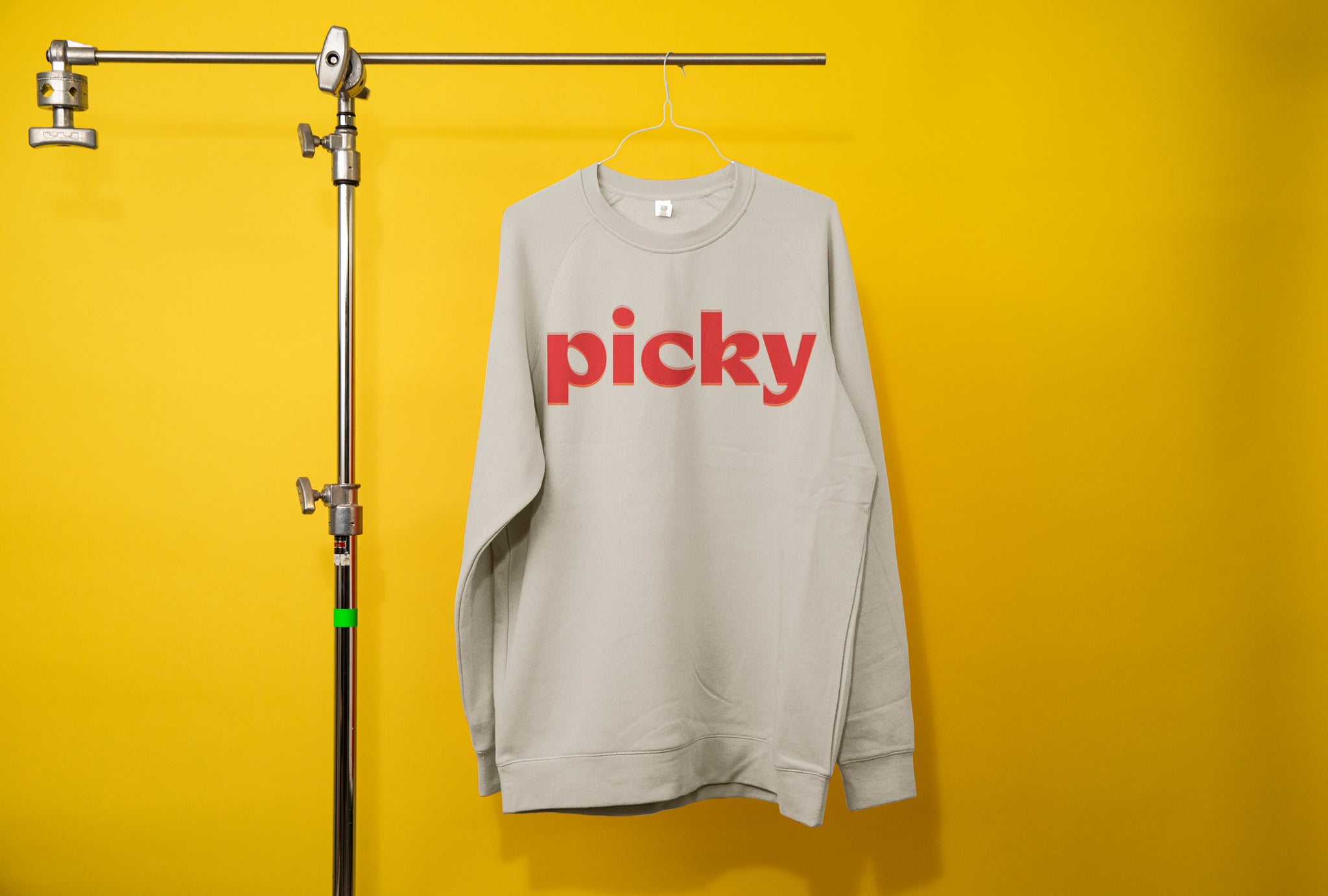 Photography c-stand holding a grey jumper. The word Picky is written in a bold red front across the chest. Background is a bright yellow. 