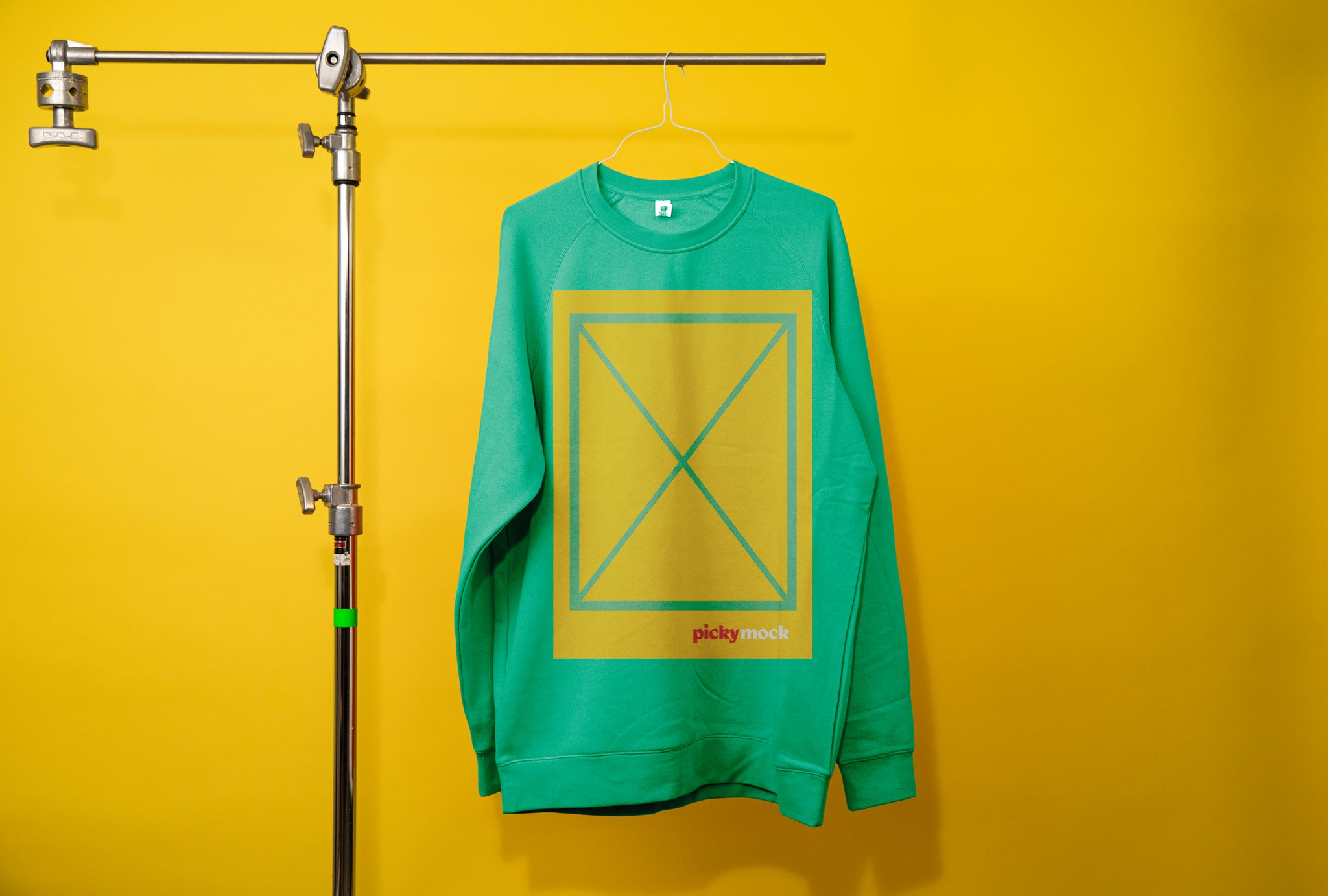 Photography c-stand holding a green jumper. Middle of jumper there is a large yellow box with green grid lines with an X in the middle. The words Picky Mock is written at the bottom of the box, just inside the line. 