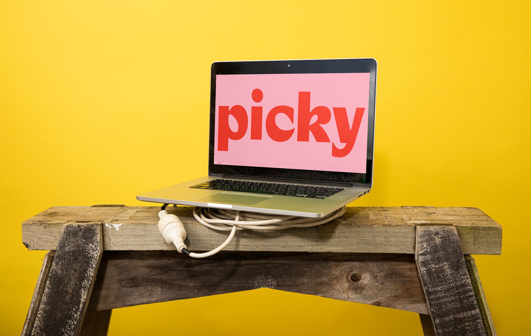Landscape close-up of a MacBook computer atop a white power cable and wooden saw horse in front of a yellow backdrop. The screen is pink with large red picky logo text.