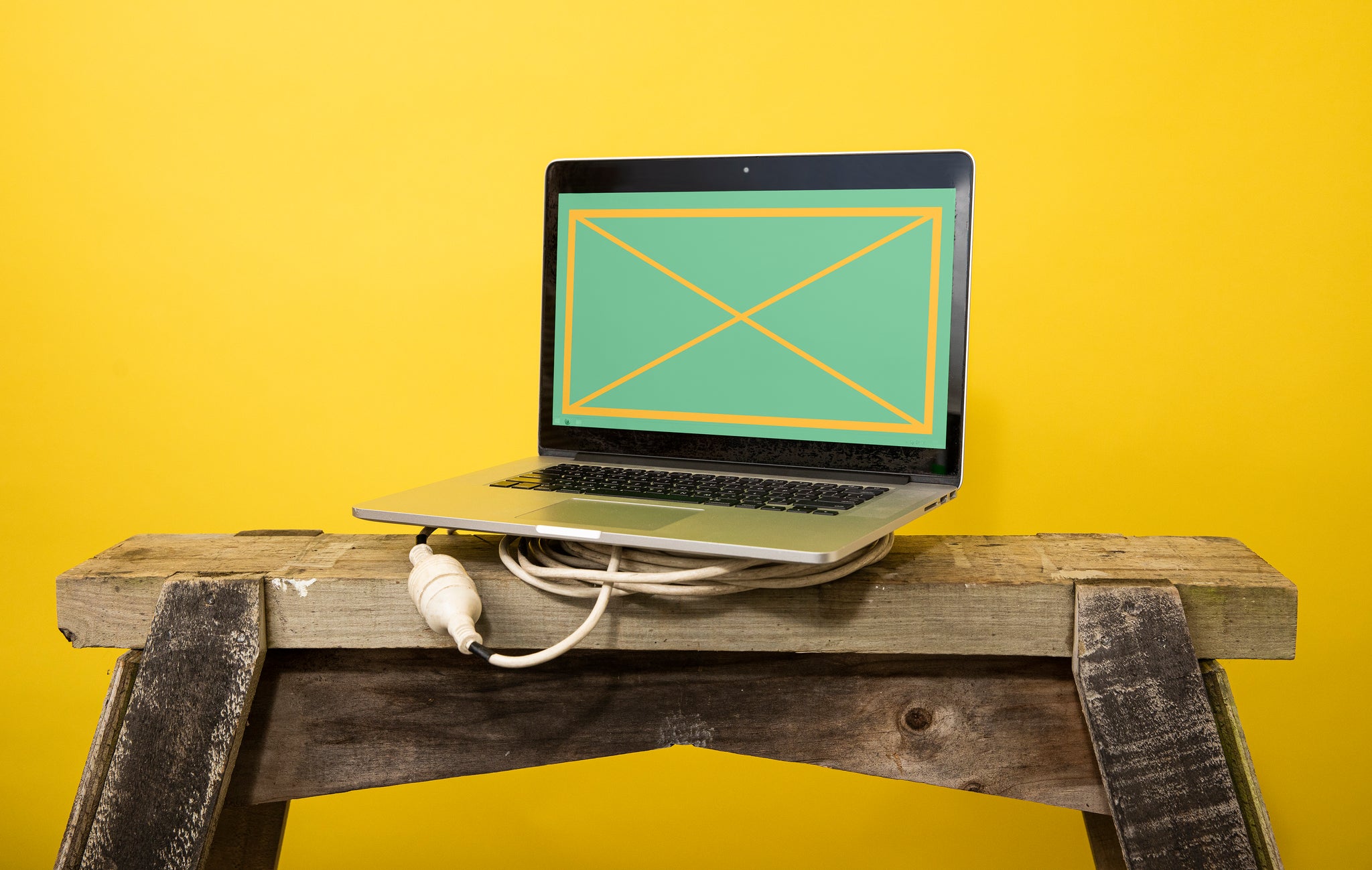 Landscape close-up of a MacBook computer atop a white power cable and wooden saw horse in front of a yellow backdrop. The screen is green and yellow.