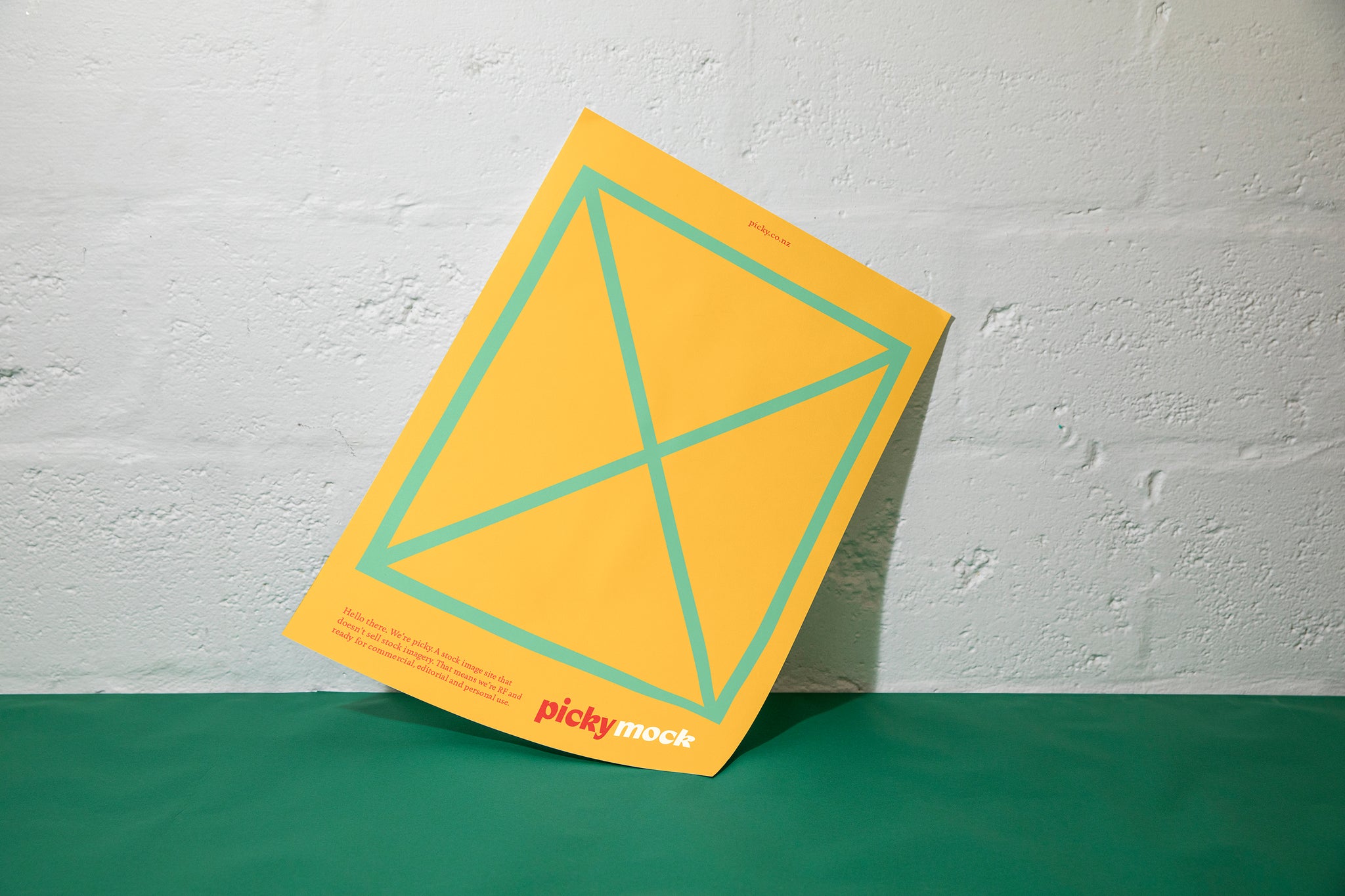 A yellow piece of card is sitting against a white cinder block wall and green surface. The card has a green box and grid lines with an X in the middle of the card. Small copy including a logo is visble at the bottom half. 