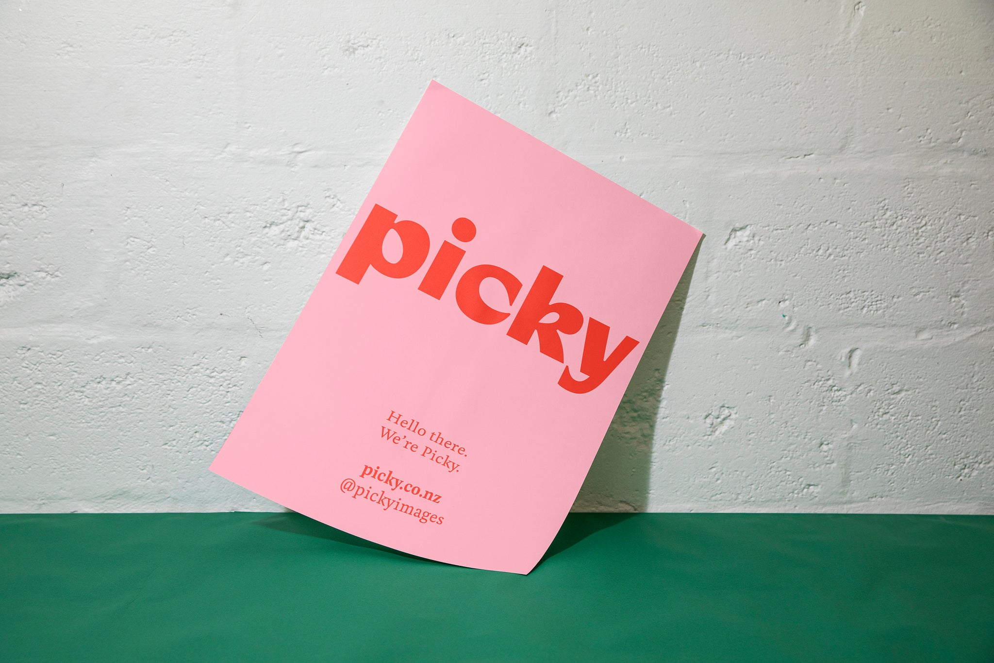 A pink piece of card is sitting against a white cinder block wall and green surface. The card is pink with the word Picky written largely cross the paper. Small copy is visble at the bottom half. 