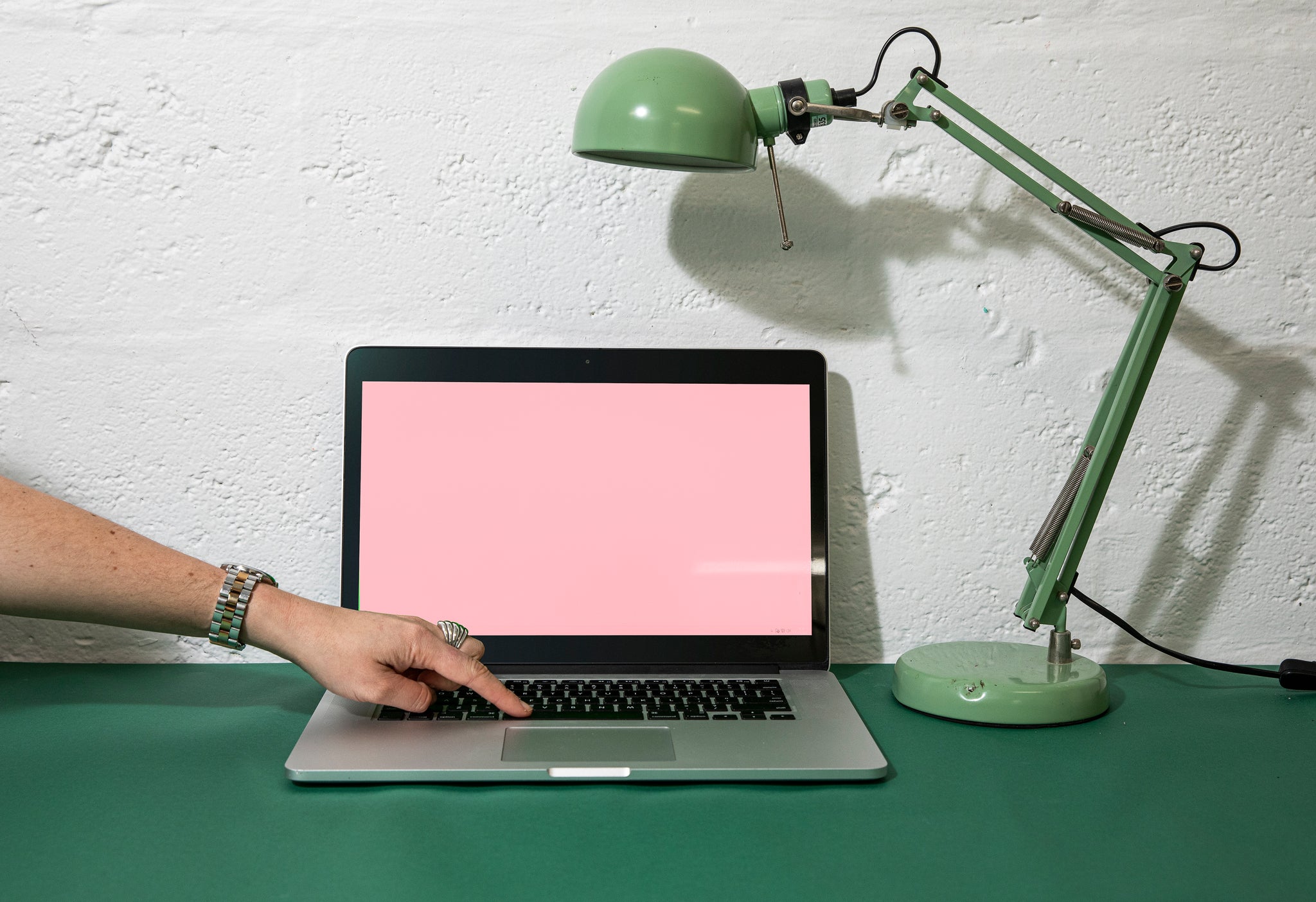 Laptop and green lamp sitting on a desk. Surface is a dark green. Laptop screen is up and a pink frame is on the screen, with a green grid line design. A womans hand is reaching into frame touching the keyboard. 
