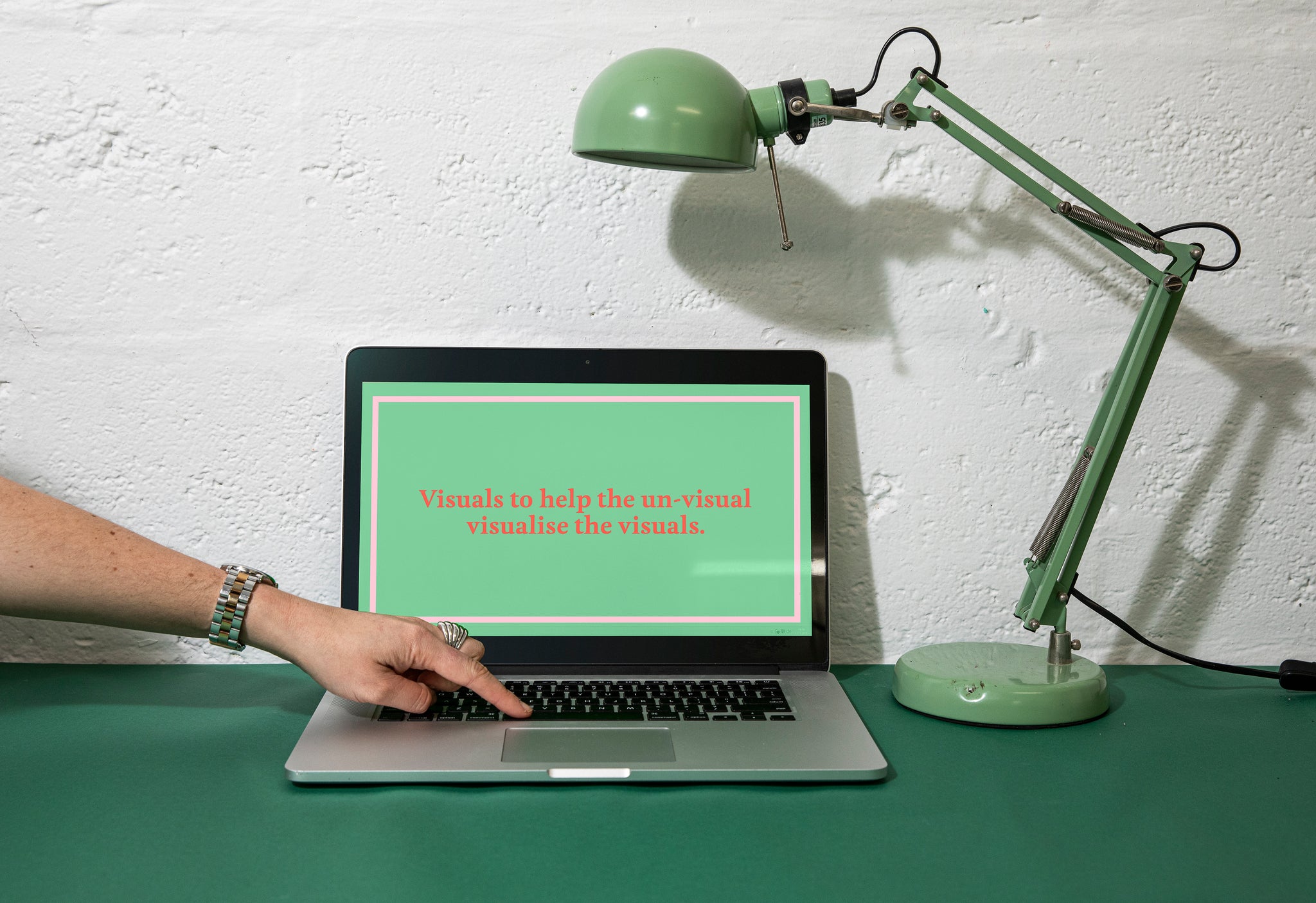 Laptop and green lamp sitting on a desk. Surface is a dark green. Laptop screen is up and a green frame is on the screen, with the words Visuals to help the un-visual visualise the visuals. A womans hand is reaching into frame touching the keyboard. 