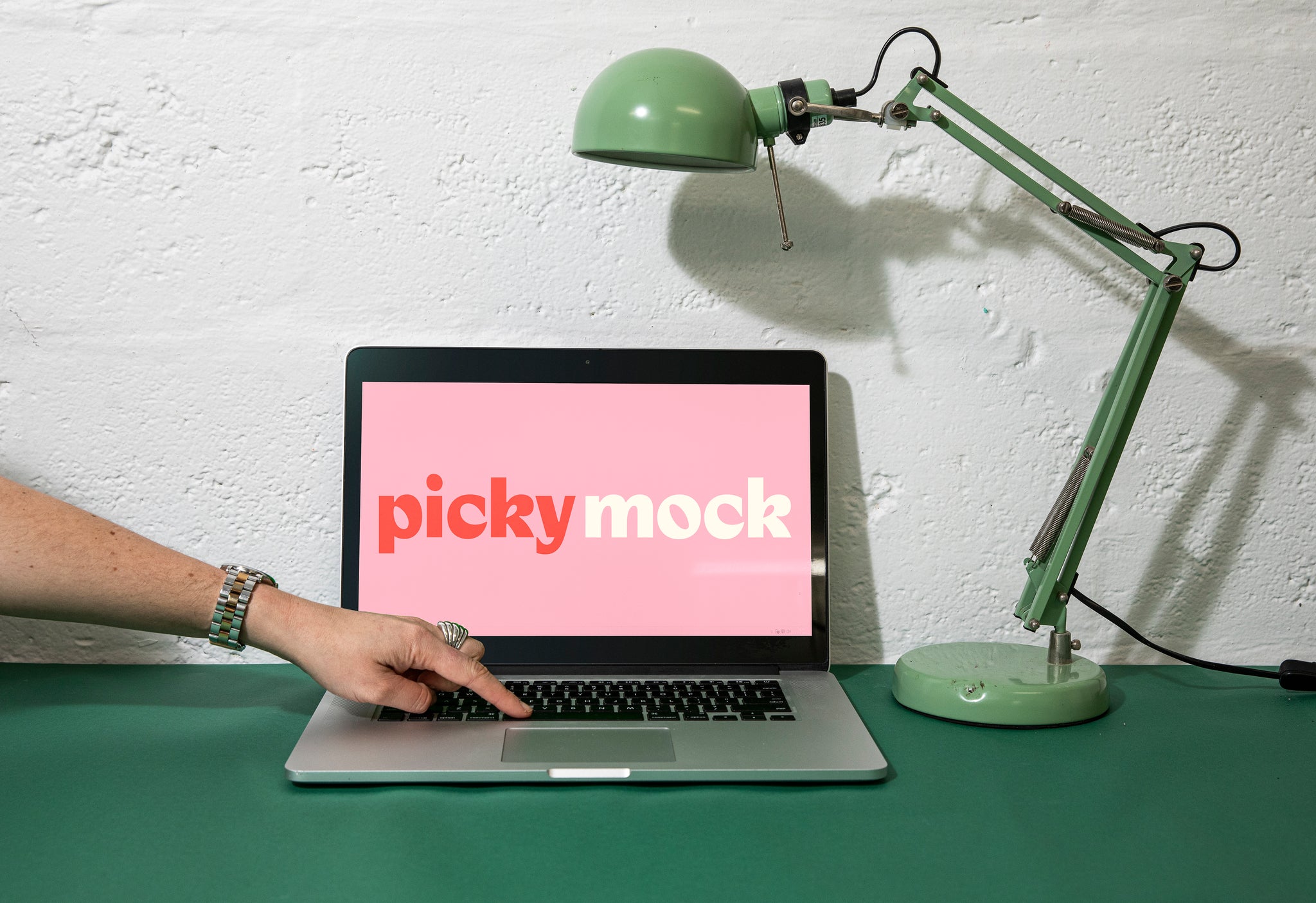 Laptop and green lamp sitting on a desk. Surface is a dark green. Laptop screen is up and a pink frame is on the screen, with the words Picky Mock written on the screen in a bold font. A womans hand is reaching into frame touching the keyboard. 