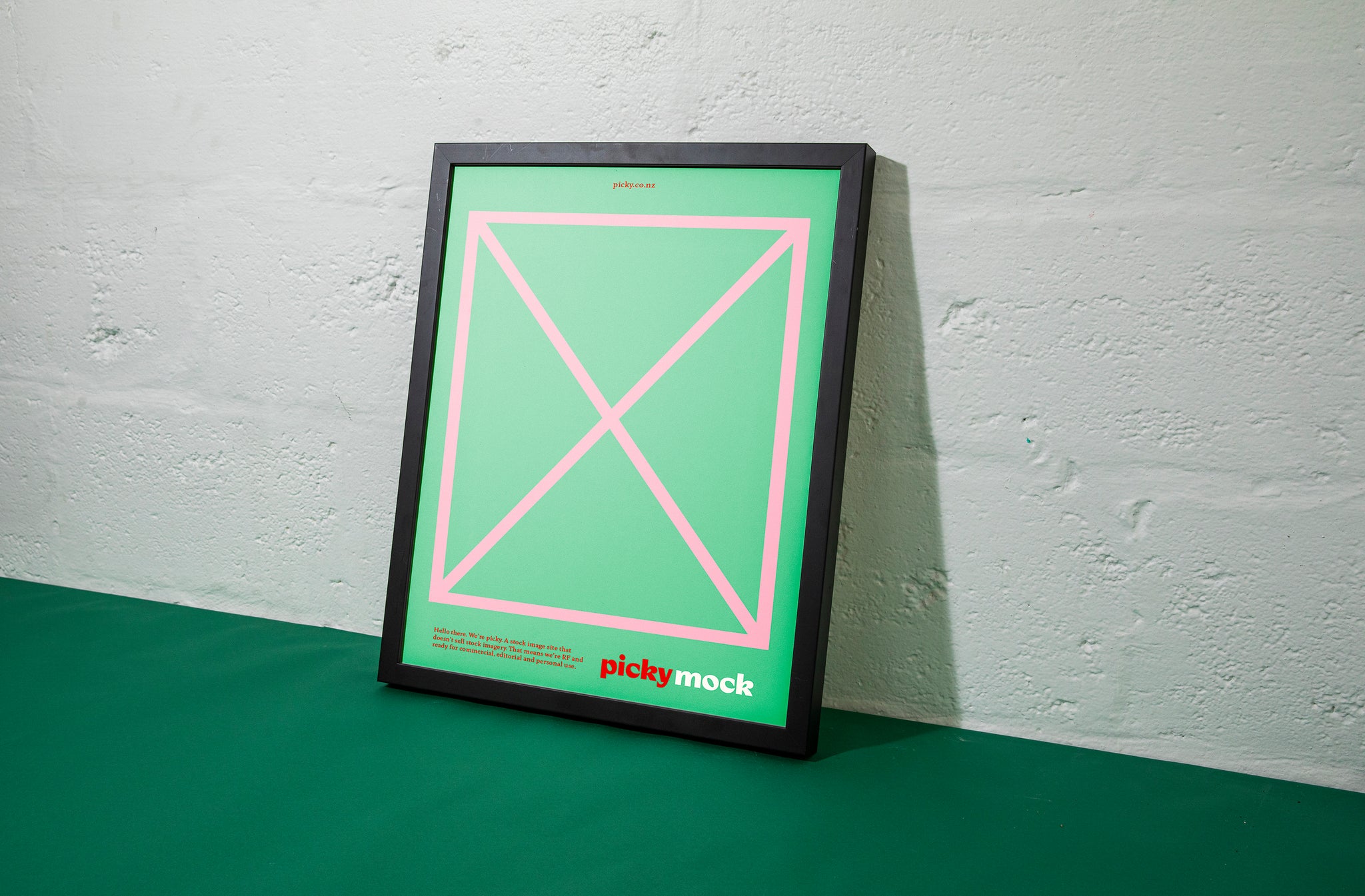 A black frame is leaning against a white cinder block wall. Inside the frame is a green card with a pink border with grid lines and X in rhe middle. Small copy including a logo is visble at the bottom half. 