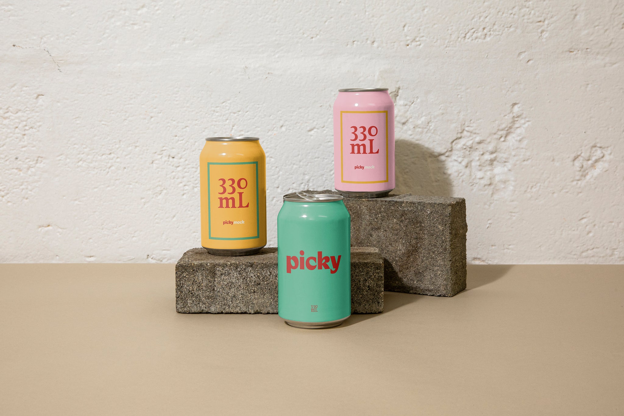Landscape close-up of three 330ml cans posed atop grey bricks at different heights in front of a white brick wall; they are pink, yellow, and green.