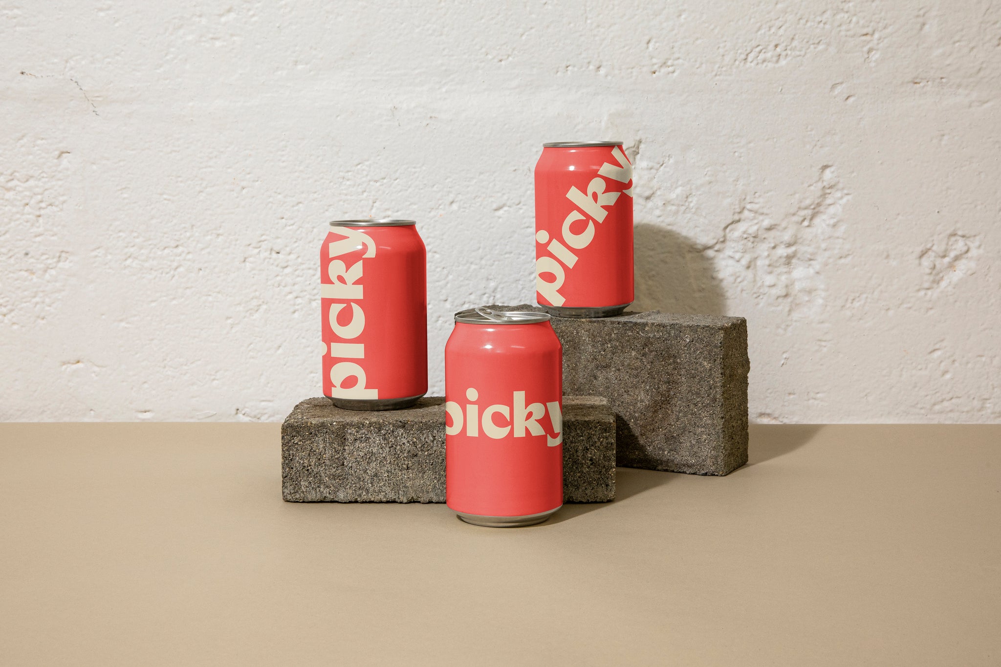 Landscape close-up of three 330ml cans posed atop grey bricks at different heights in front of a white brick wall; they are red with large white picky logo across the face.