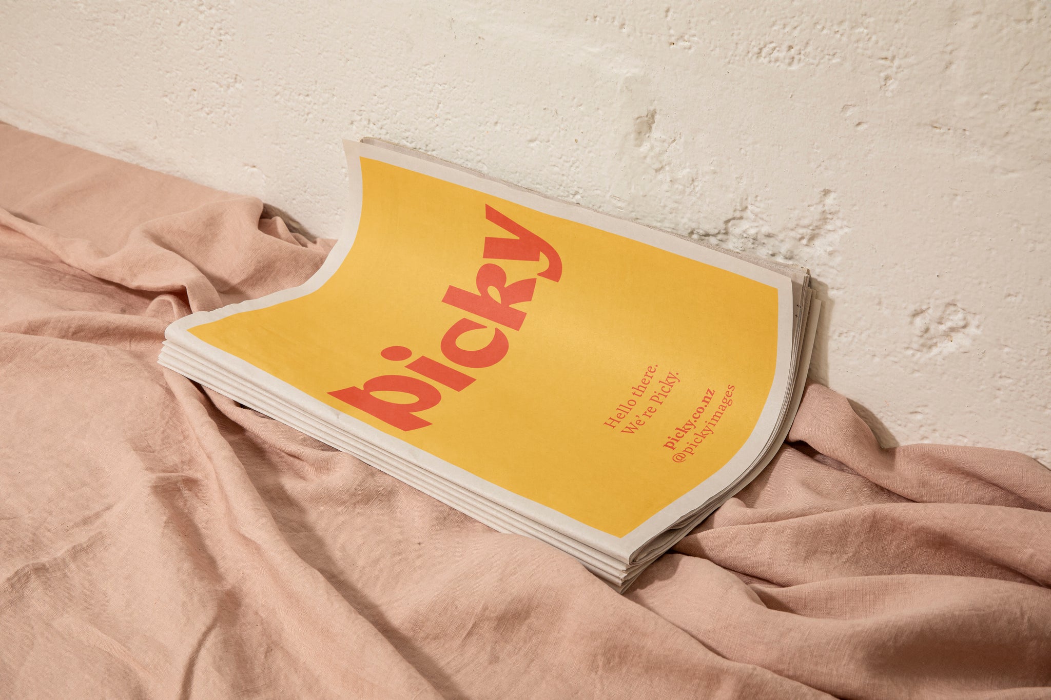 Mockup of a curved tabloid-sized newspaper sitting against a white concrete wall. A light pink cloth sits ruffled underneath.