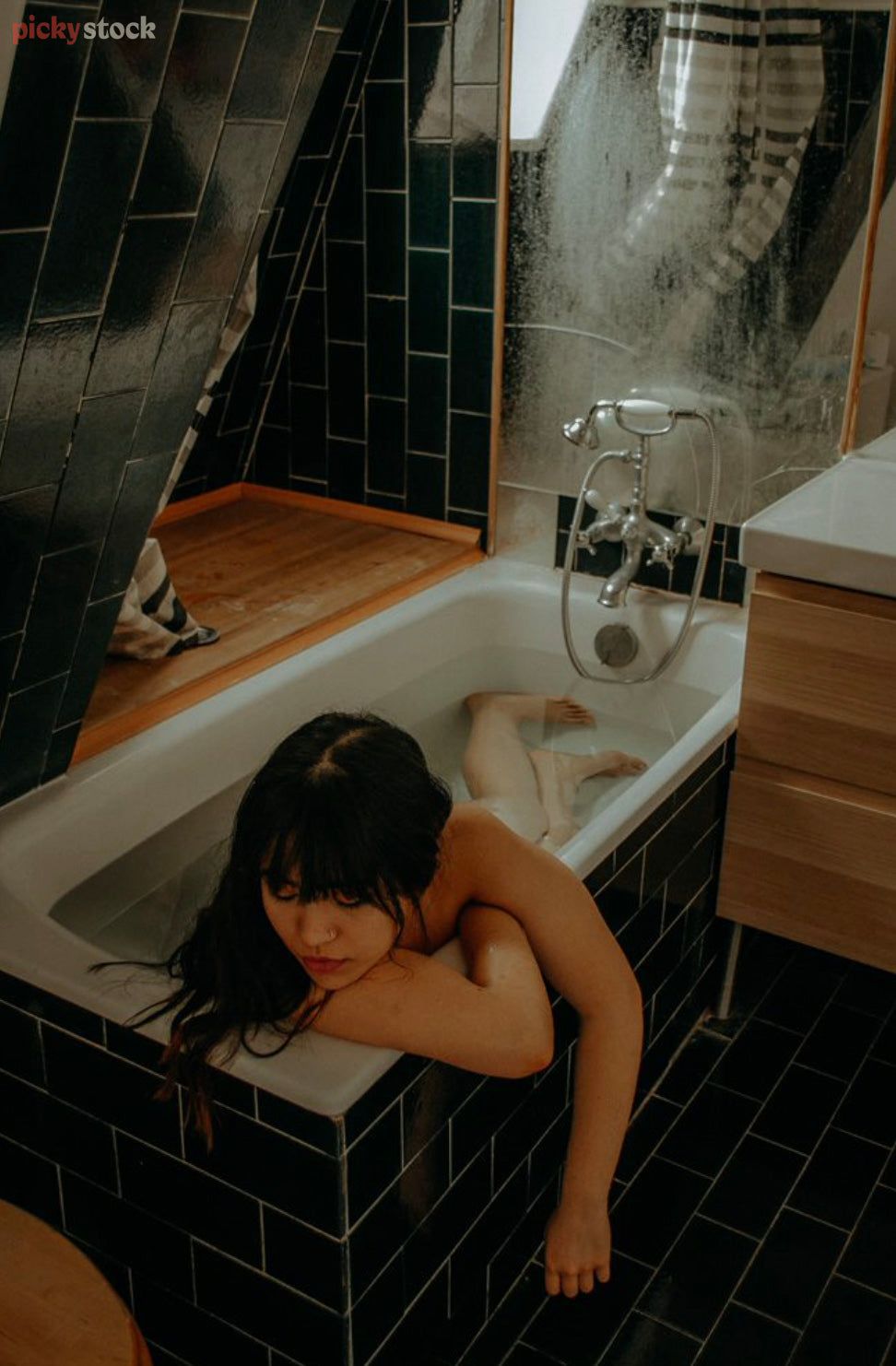 A lady lies in a bathtub in a dark tiled bathroom. She artfully covers herself with arms leaning on the edge of the bath. 