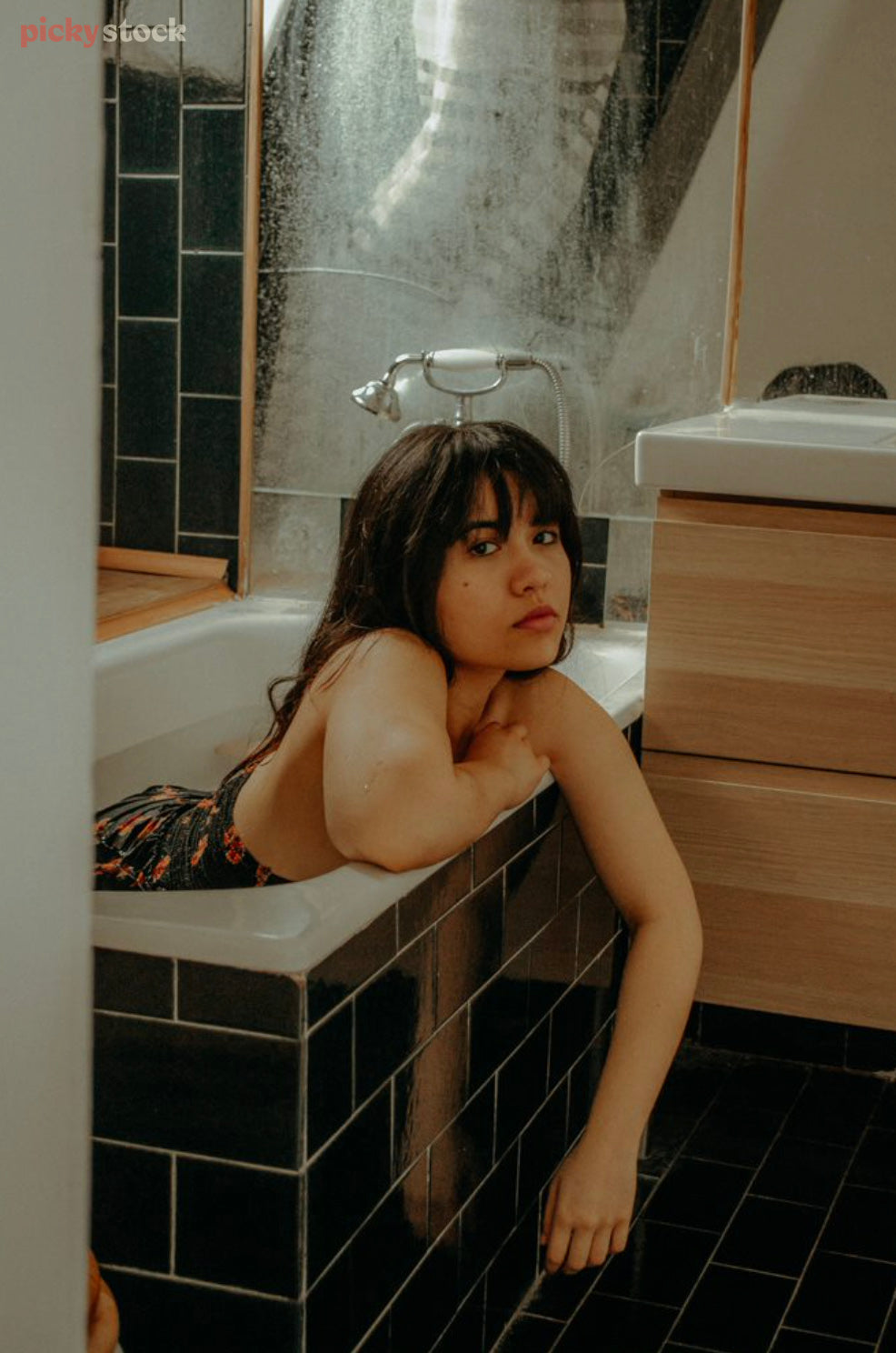 A lady lies peers out to camera from a bathtub in a dark tiled bathroom. She artfully covers herself with arms leaning on the edge of the bath as she gazes direct to camera. 
