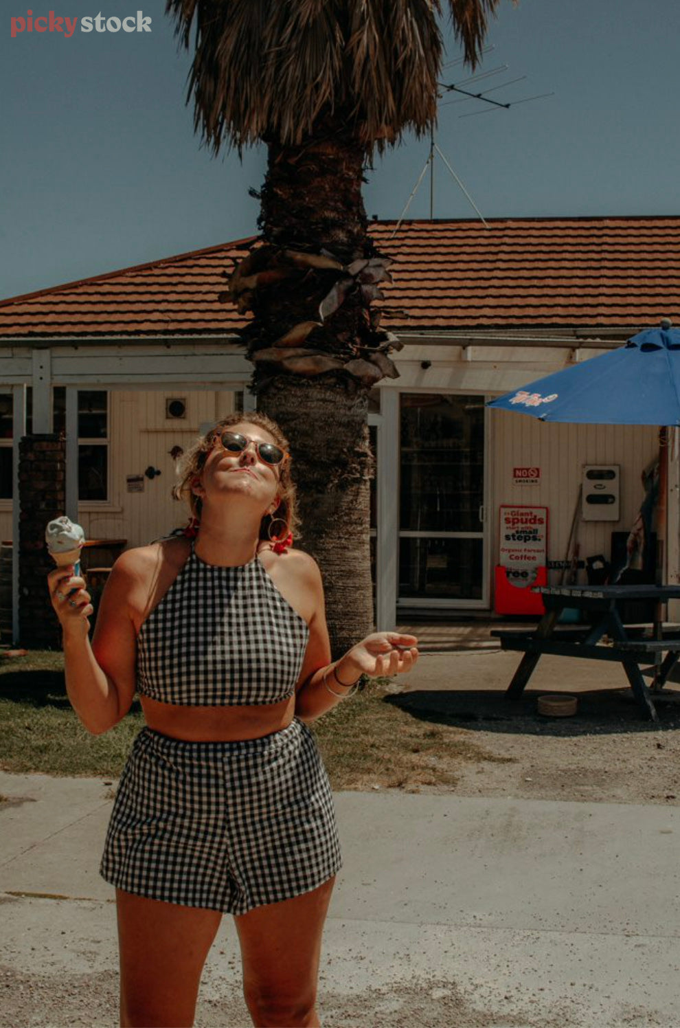 A young lady in a match-ing two piece gingham outfit pulls a funny face to the camera as he eats her classic Kiwi ice-cream on a hot summers day. She stands outside the ice cream shop under an old large palm tree.