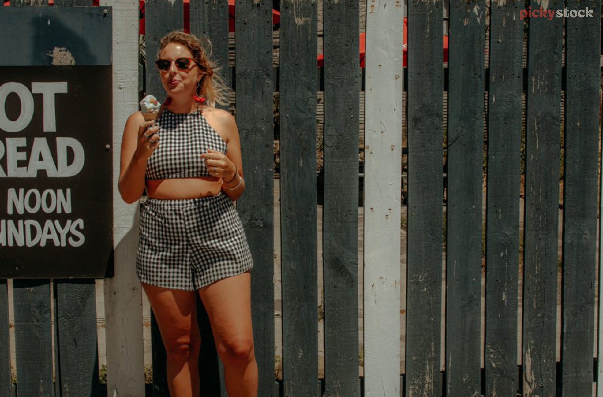 A young lady in a match-ing two piece gingham outfit eats a classic New Zealand ice-cream while standing against a characterful green and white wooden fence. A handwritten "Hot Bread" retro sign sits in frame next to her. 