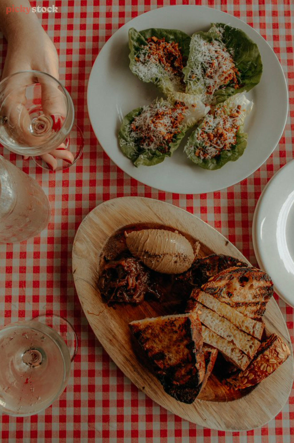 Birds-eye-view image of a plated up meal at a New Zealand restaurant. The restaurant food sits on a red and white checked tablecloth, with a hand holding a glass of white wine. 