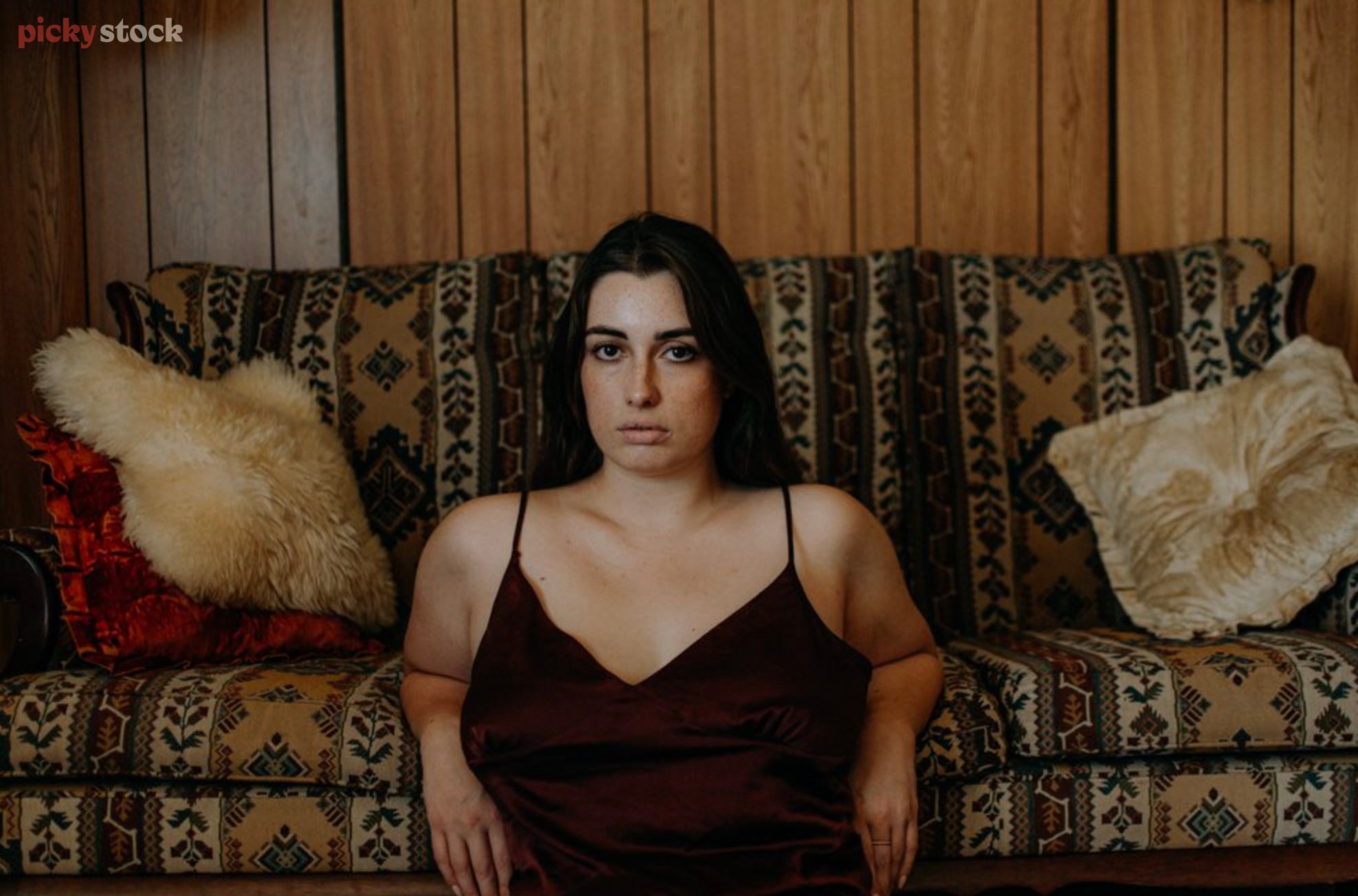 A brunette lady in her twenties gazes directly to camera as she leans on the eighties style retro patterned couch in a New Zealand house. 
